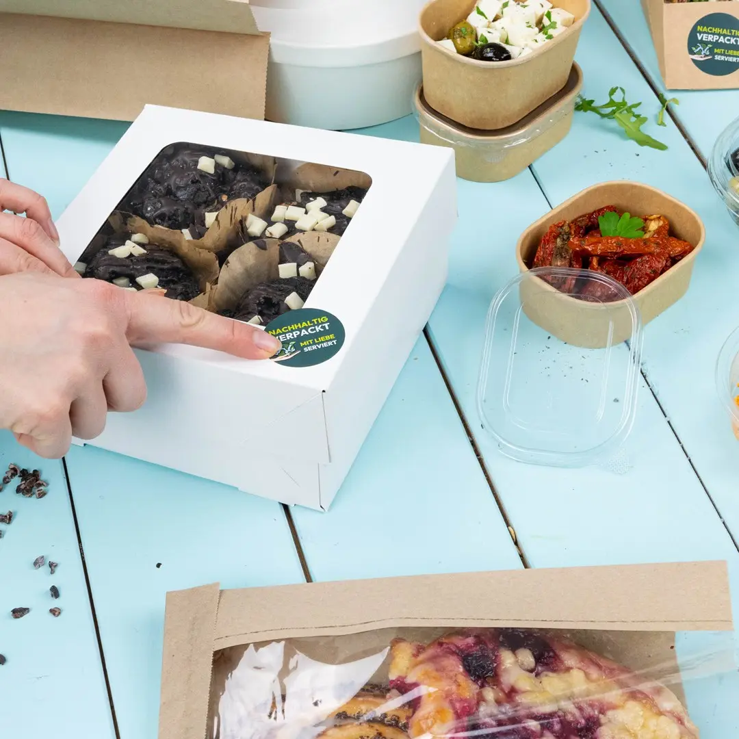 Scatola bianca contenente prodotti da forno con coperchio trasparente, circondata da vari contenitori alimentari su un tavolo blu.
