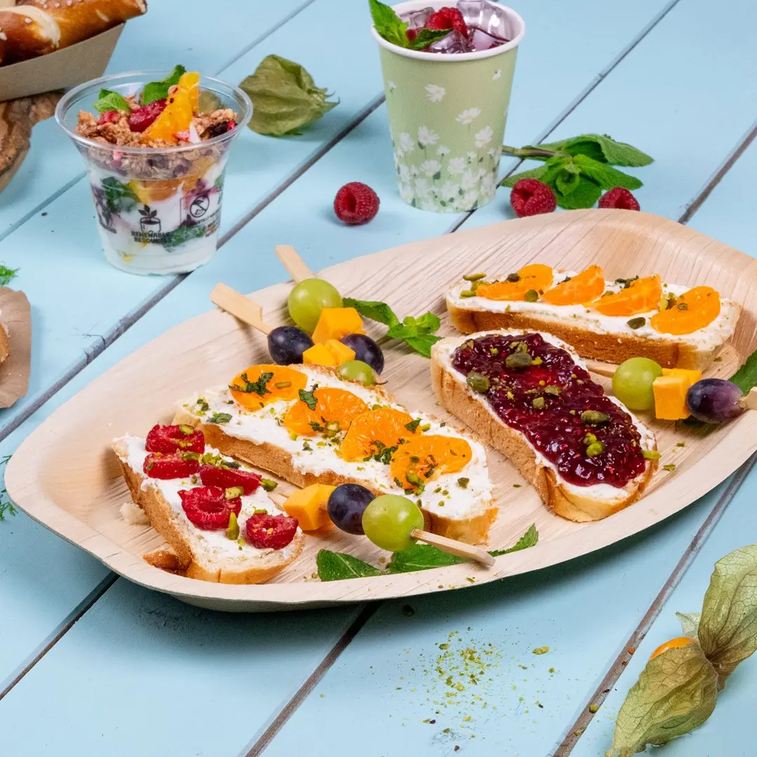 Vassoio con vari finger food tra cui pane con formaggio cremoso, frutta e uno spiedino di frutta, insieme a un parfait di yogurt.
