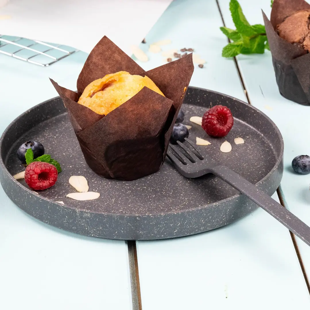 Muffin in un involucro di carta marrone su un piatto grigio con lamponi, mirtilli, fette di mandorle e una piccola forchetta.