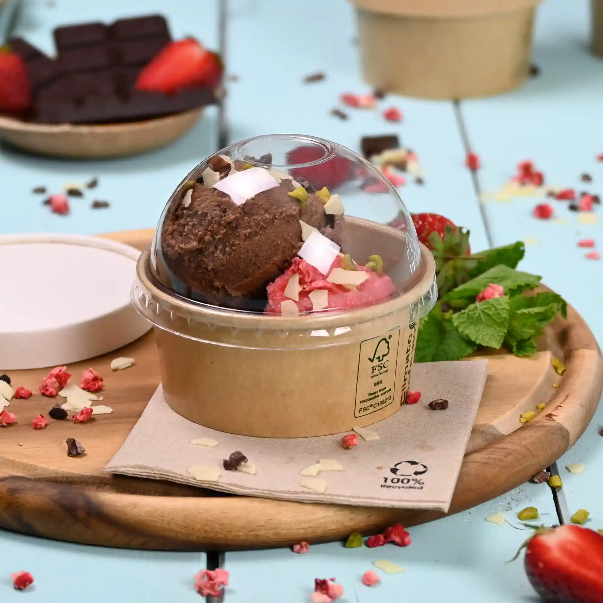 Contenitore con due palline di gelato, guarnito con noci e cocco, su un tagliere di legno con menta e fragole.