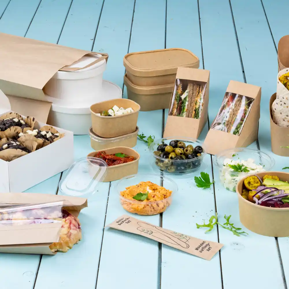 Un assortimento di contenitori per alimenti include scatole di carta, ciotole e bicchieri di plastica trasparente con vari piatti e snack.