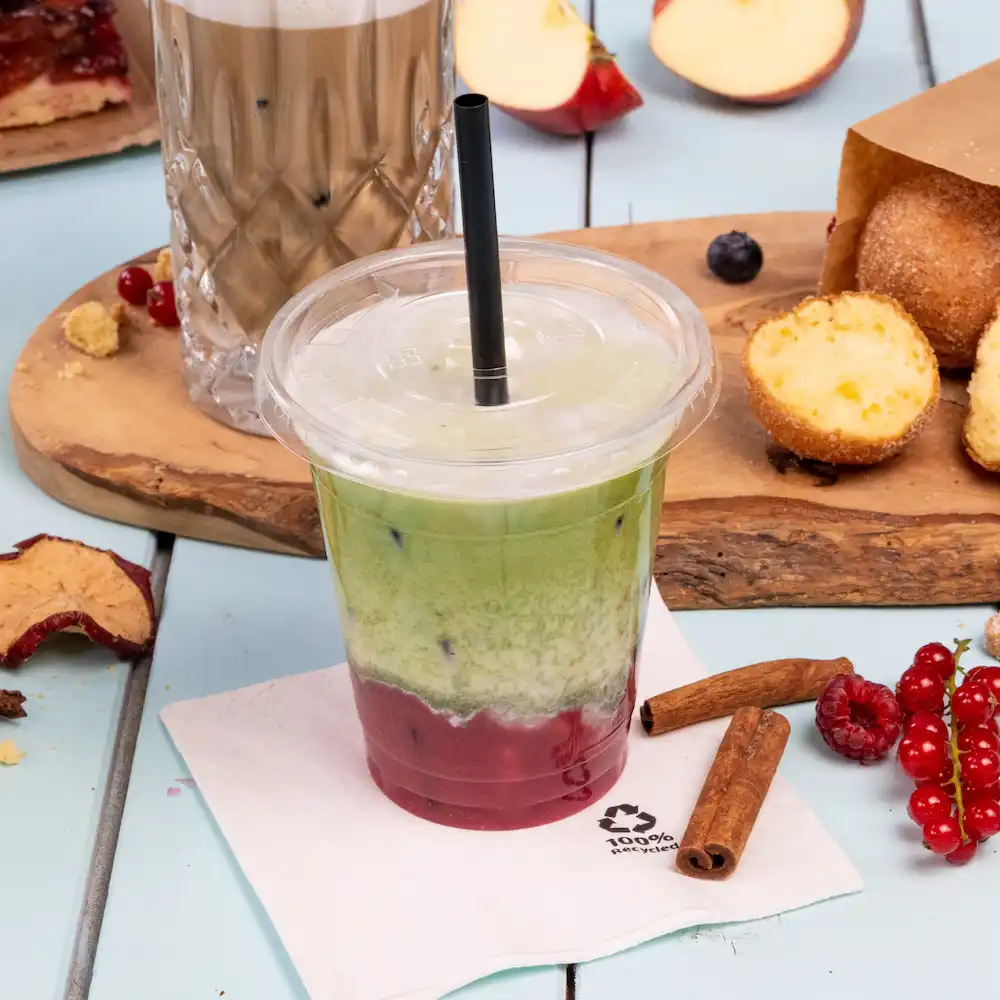 Bevanda ghiacciata a strati in un bicchiere trasparente con una cannuccia, circondata da snack e un tovagliolo su una superficie azzurro chiaro.