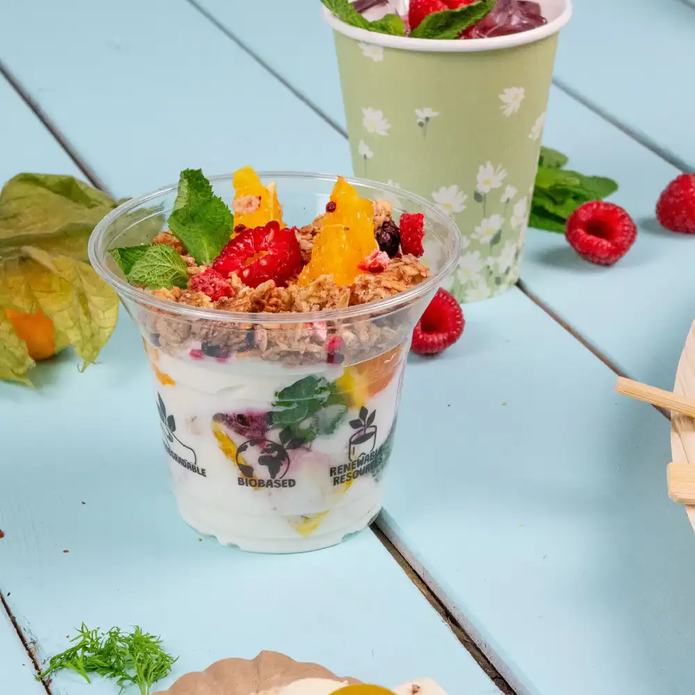 Bicchiere di dessert in plastica trasparente riempito di yogurt, frutta e granola, posizionato su una superficie blu con foglie di menta vicine.