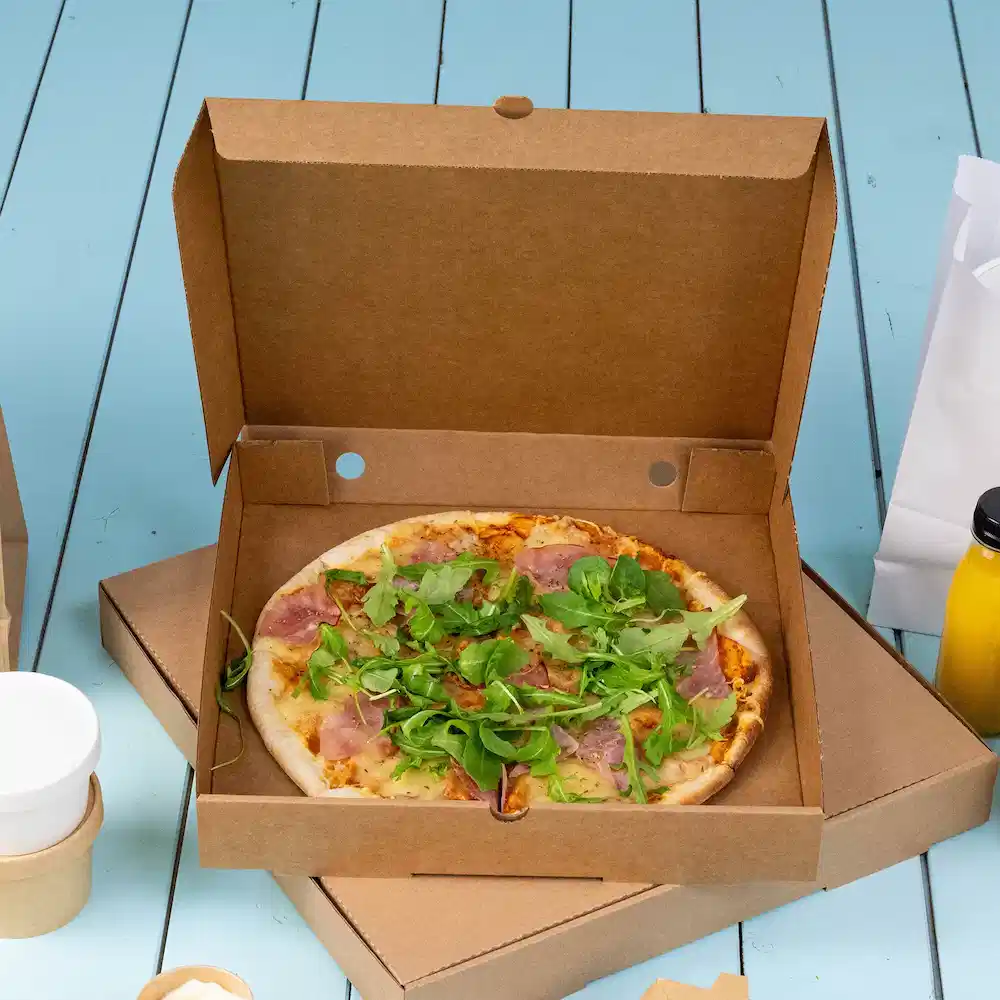 Pizza in una scatola da asporto di cartone, con prosciutto e misticanza su una superficie in legno blu.