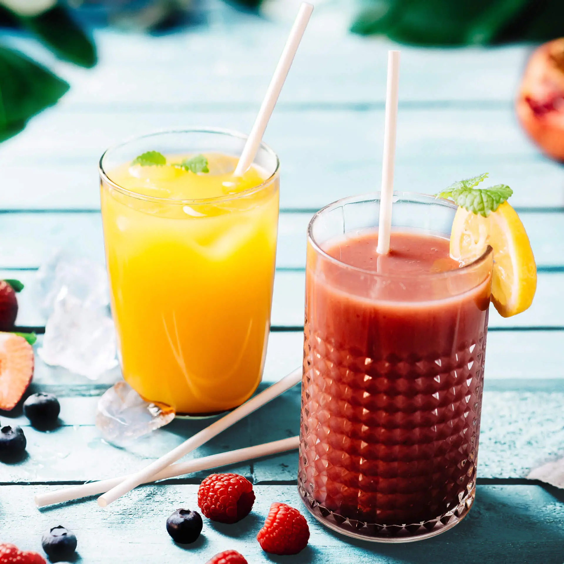 Due bicchieri di bevande fredde su un tavolo di legno: uno con bevanda gialla e ghiaccio, l'altro con bevanda rossastra guarnita di limone. Cannucce bianche biodegradabili e frutti di bosco freschi sparsi.