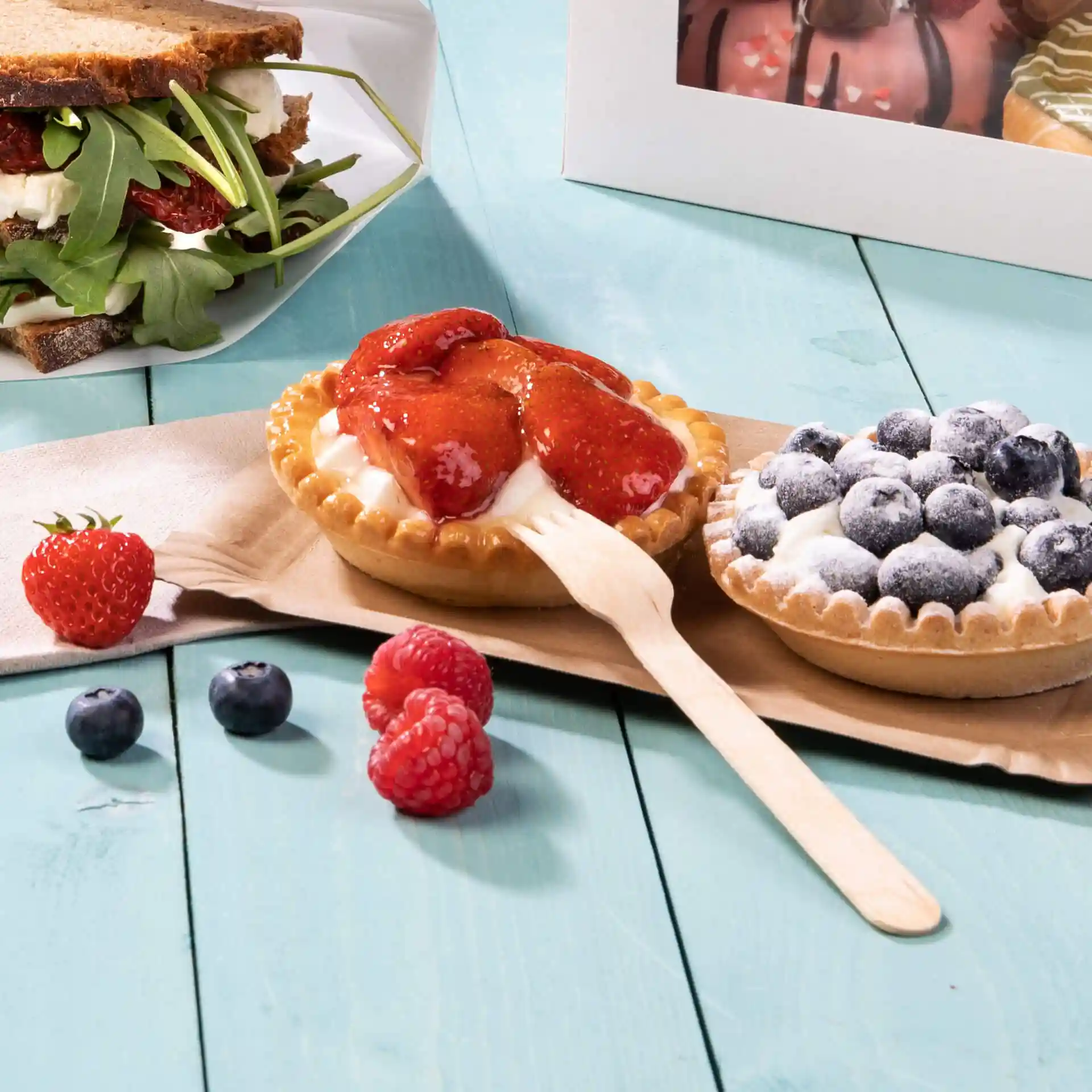 Immagine di due crostate dessert con fragole e mirtilli, una forchetta di legno e frutti di bosco freschi su una superficie di legno blu.