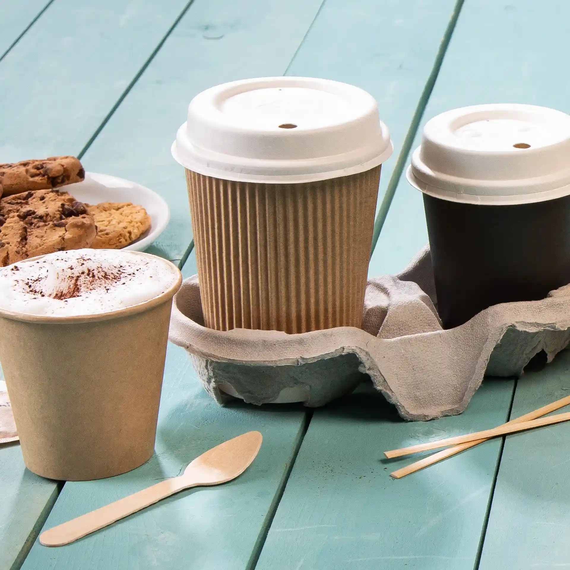 Immagine di una tazza di caffè marrone a righe, una tazza di caffè bianca e un portabicchieri marrone con un cucchiaio di legno e biscotti su una superficie turchese.