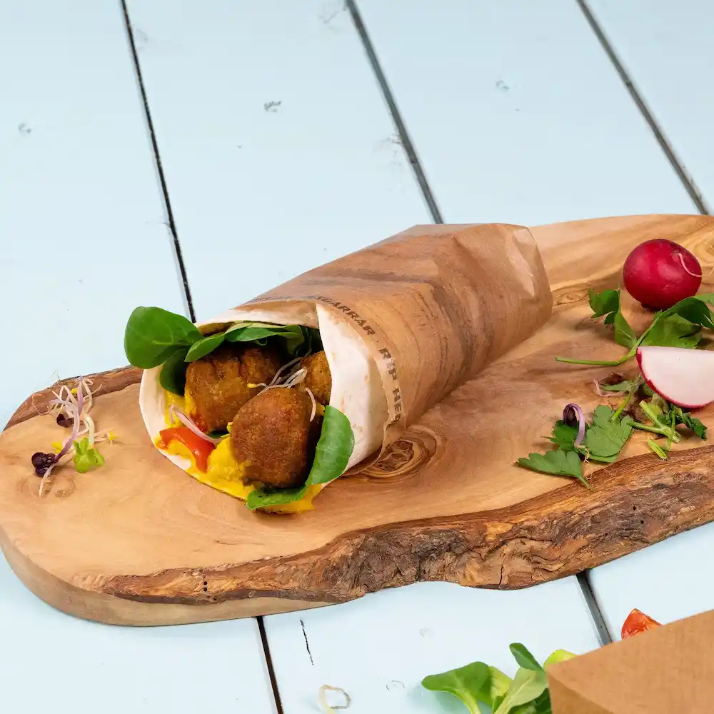 Uno snack avvolto ripieno di falafel, spinaci e salsa su un tagliere di legno, accompagnato da ravanelli e erbe.