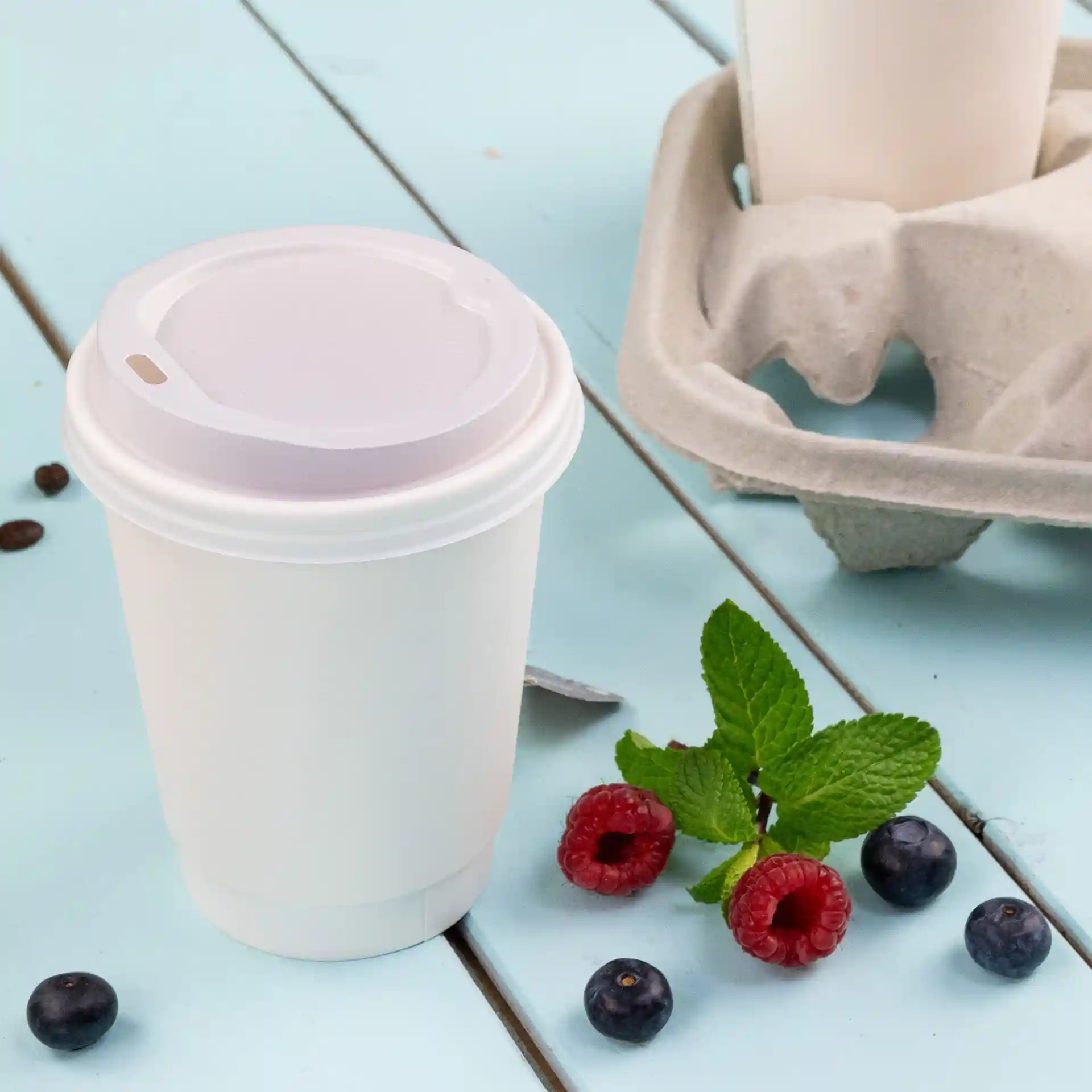 Tazza da caffè bianca a doppia parete con coperchio, 200 ml, posata su una superficie blu con lamponi, mirtilli e foglie di menta.