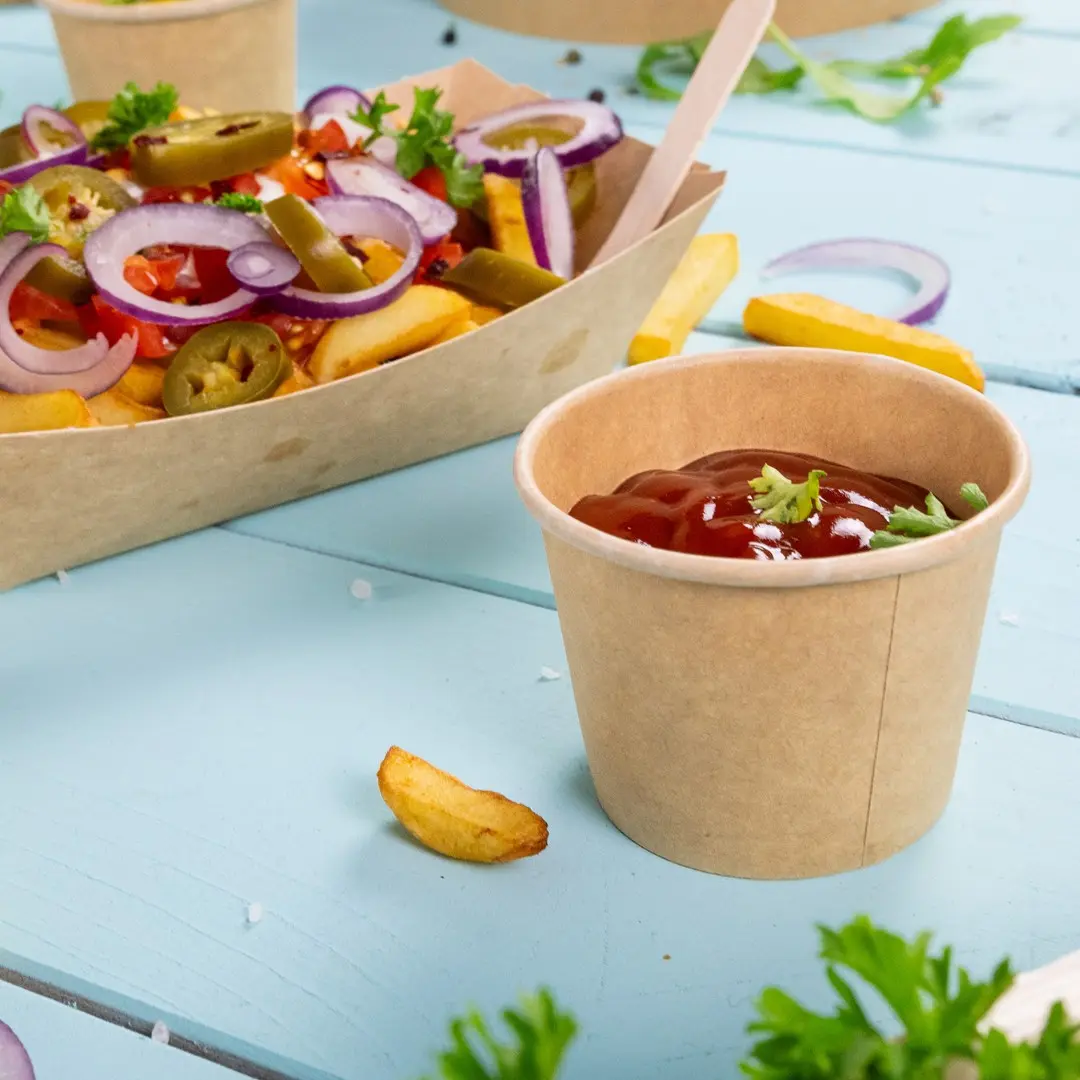 Bicchiere di carta marrone riempito di ketchup, guarnito con un piccolo pezzo di prezzemolo, accanto a una porzione di patatine e verdure.