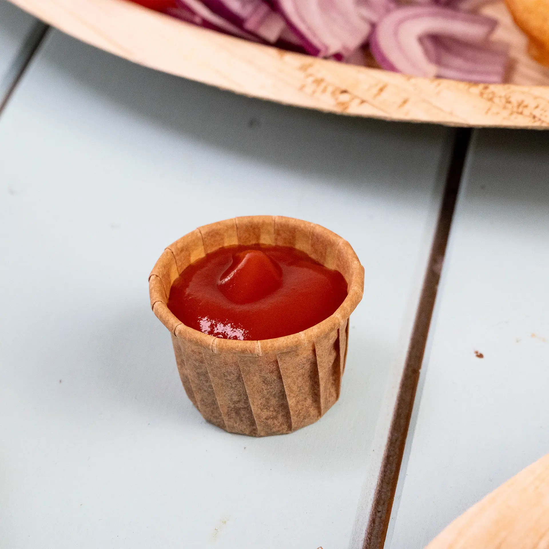 Coppetta di carta marrone da 15 ml riempita di ketchup, posizionata su una superficie blu, con cipolle affettate e un piatto sullo sfondo.