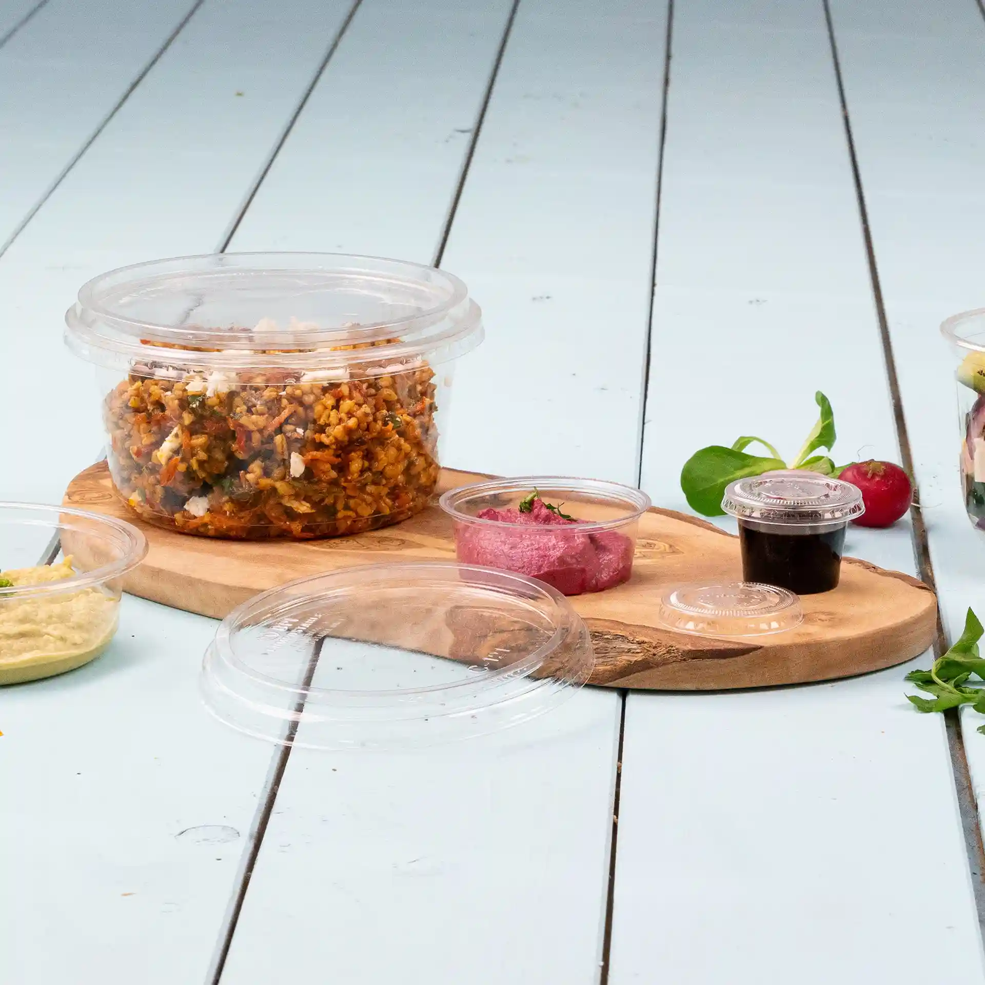 Contenitore rotondo trasparente con coperchio, riempito di cibo, su un tagliere di legno con piccoli bicchieri di salsa vicini.