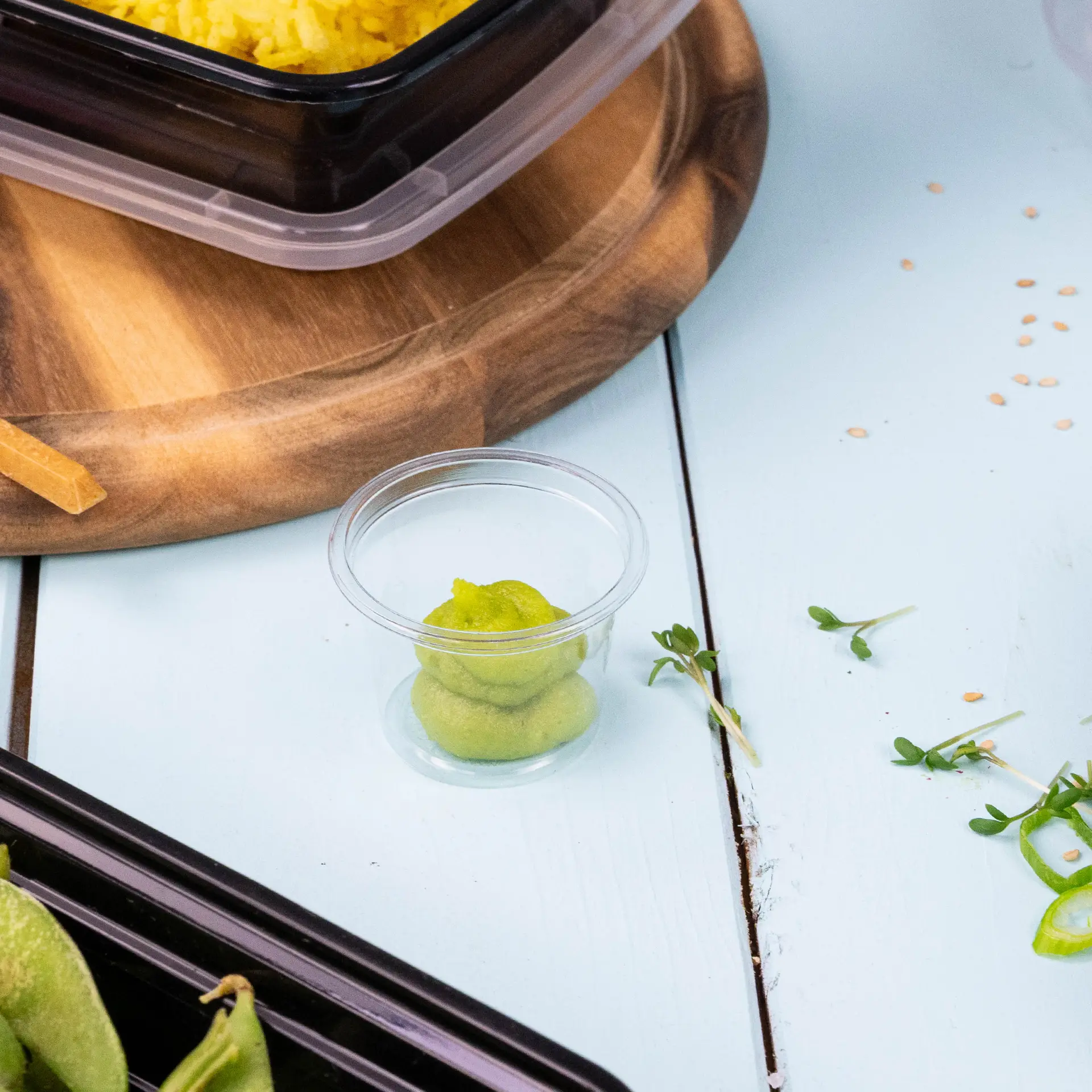 Un bicchiere di plastica trasparente con wasabi verde all'interno, posato su una superficie in legno blu chiaro vicino a imballaggi alimentari e guarnizioni.
