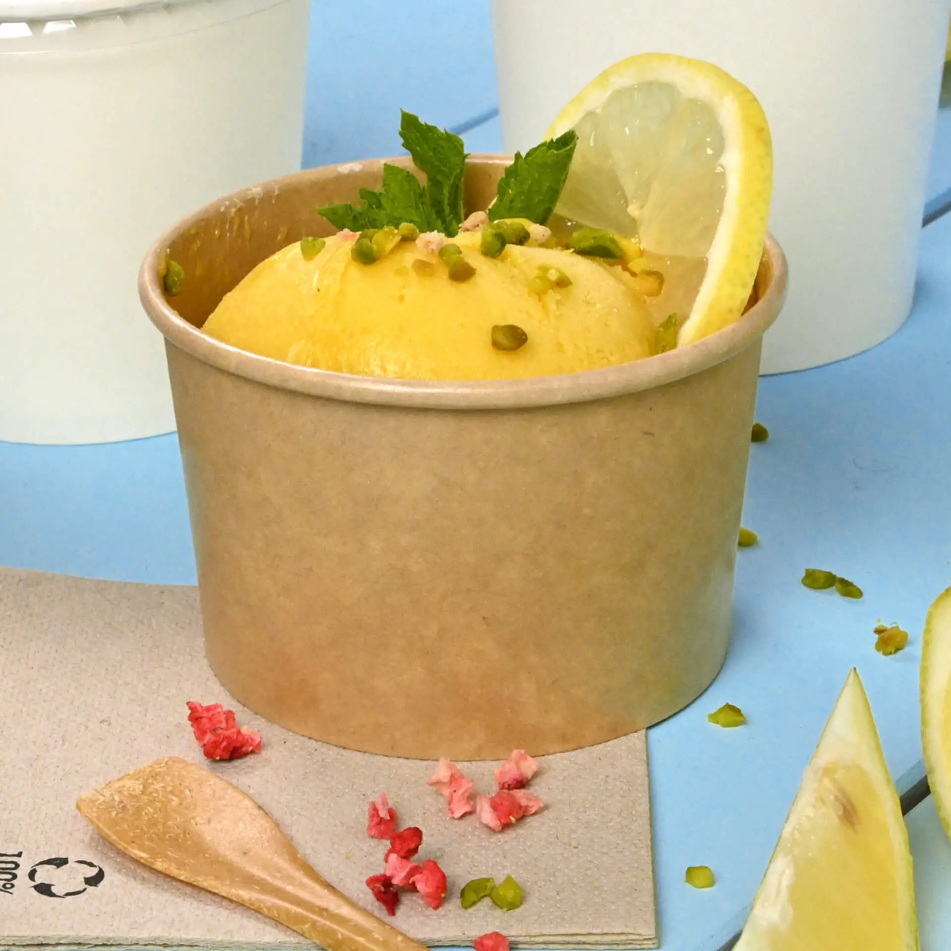 Un cono di gelato marrone riempito di gelato giallo, guarnito con menta e una fetta di limone; esposto con cucchiai e condimenti nelle vicinanze.