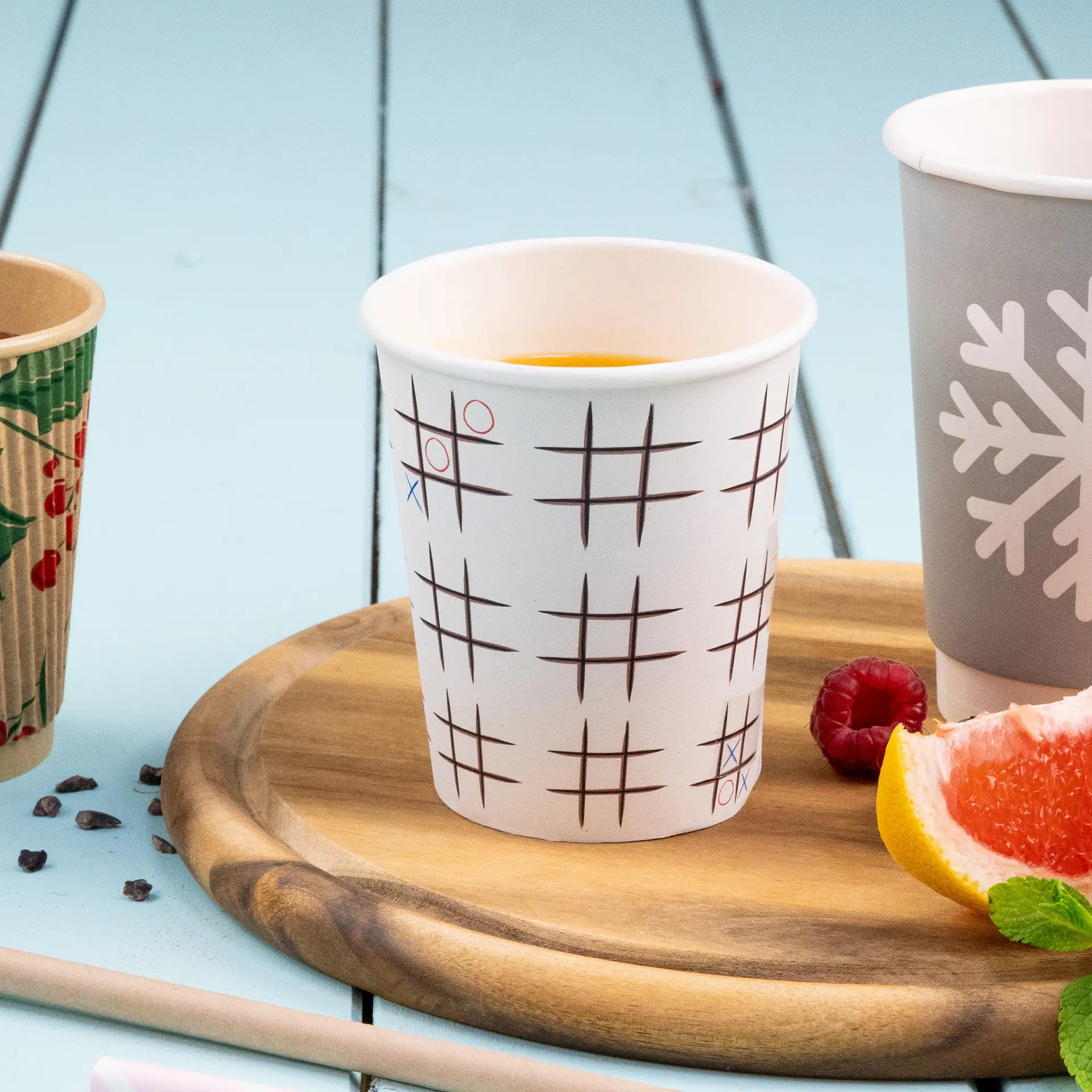 Un bicchiere di carta bianco con stampa Tic Tac Toe, contenente una bevanda; altri bicchieri simili con opacità ridotta e fette di frutta nelle vicinanze.