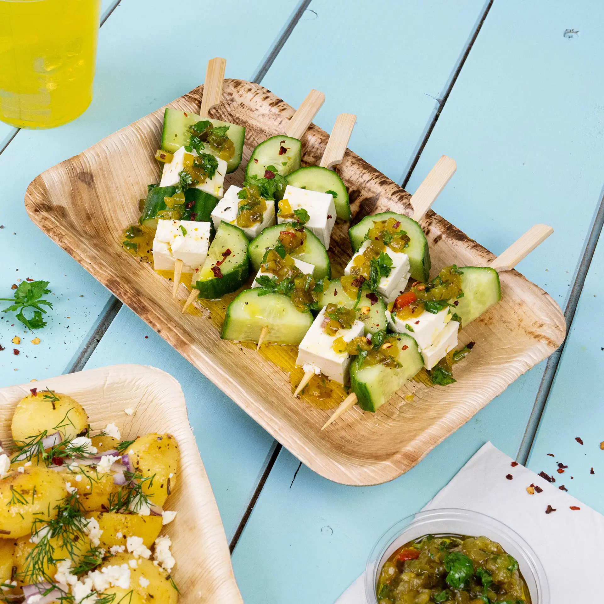 Piatto in bambù con spiedini di cetriolo e formaggio, guarnito con erbe e salsa, servito con insalata di patate.