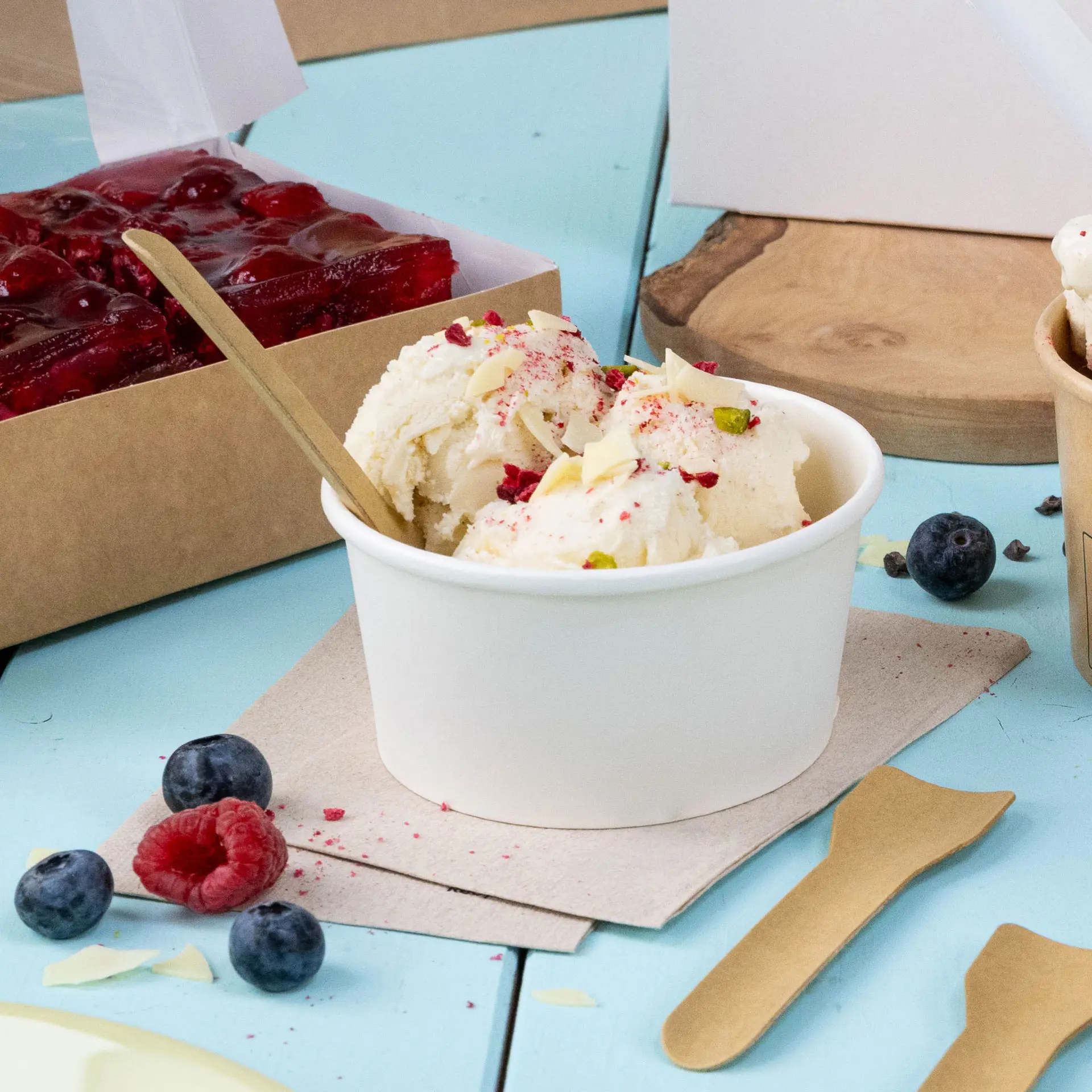 Becher di gelato guarnito con frutta e noci, cucchiaio di legno e frutti di bosco freschi su una superficie blu con un tovagliolo.