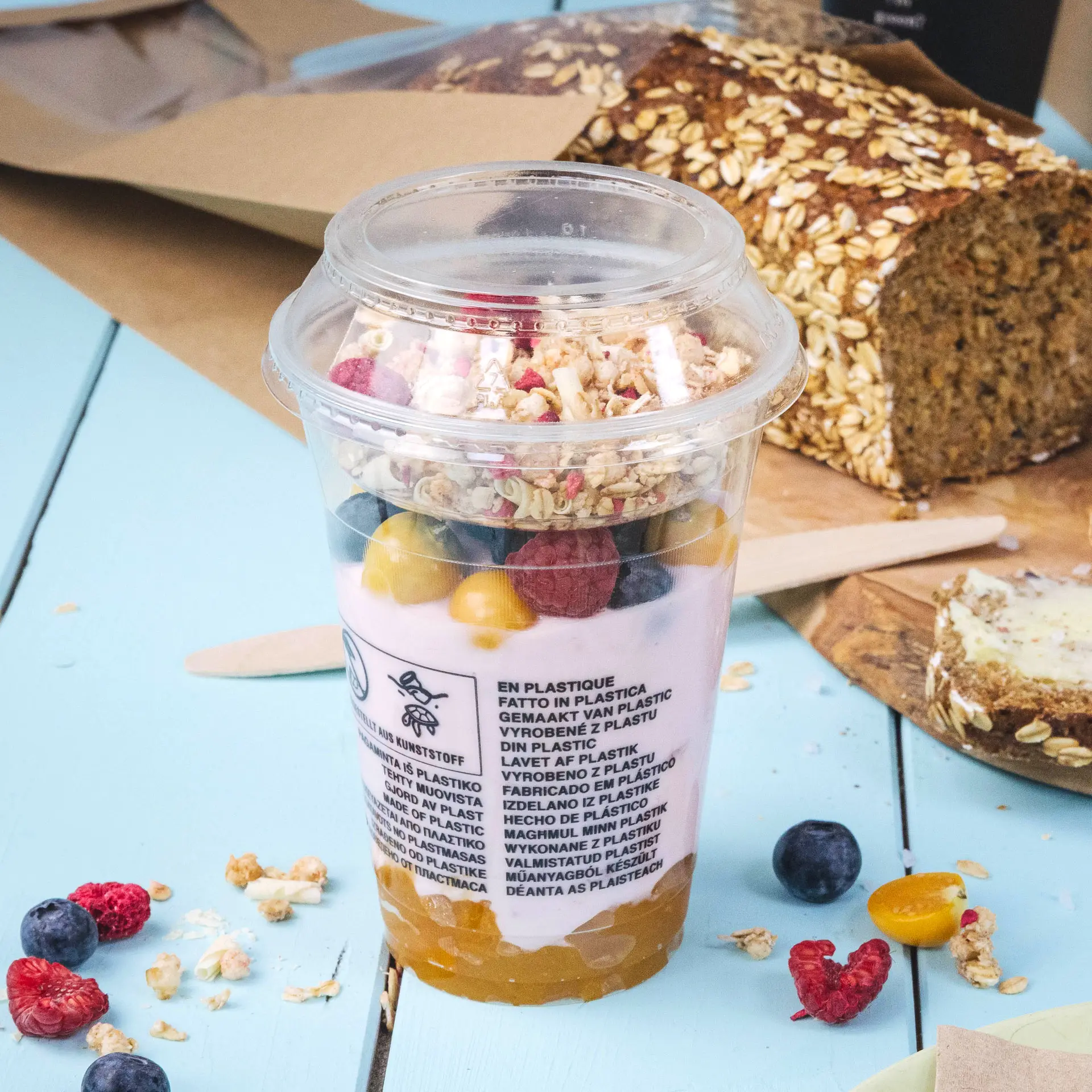 Un cup smoothie PLA trasparente da 300 ml, riempito di yogurt, frutta e granola, posato su un tavolo blu chiaro con un pane accanto.