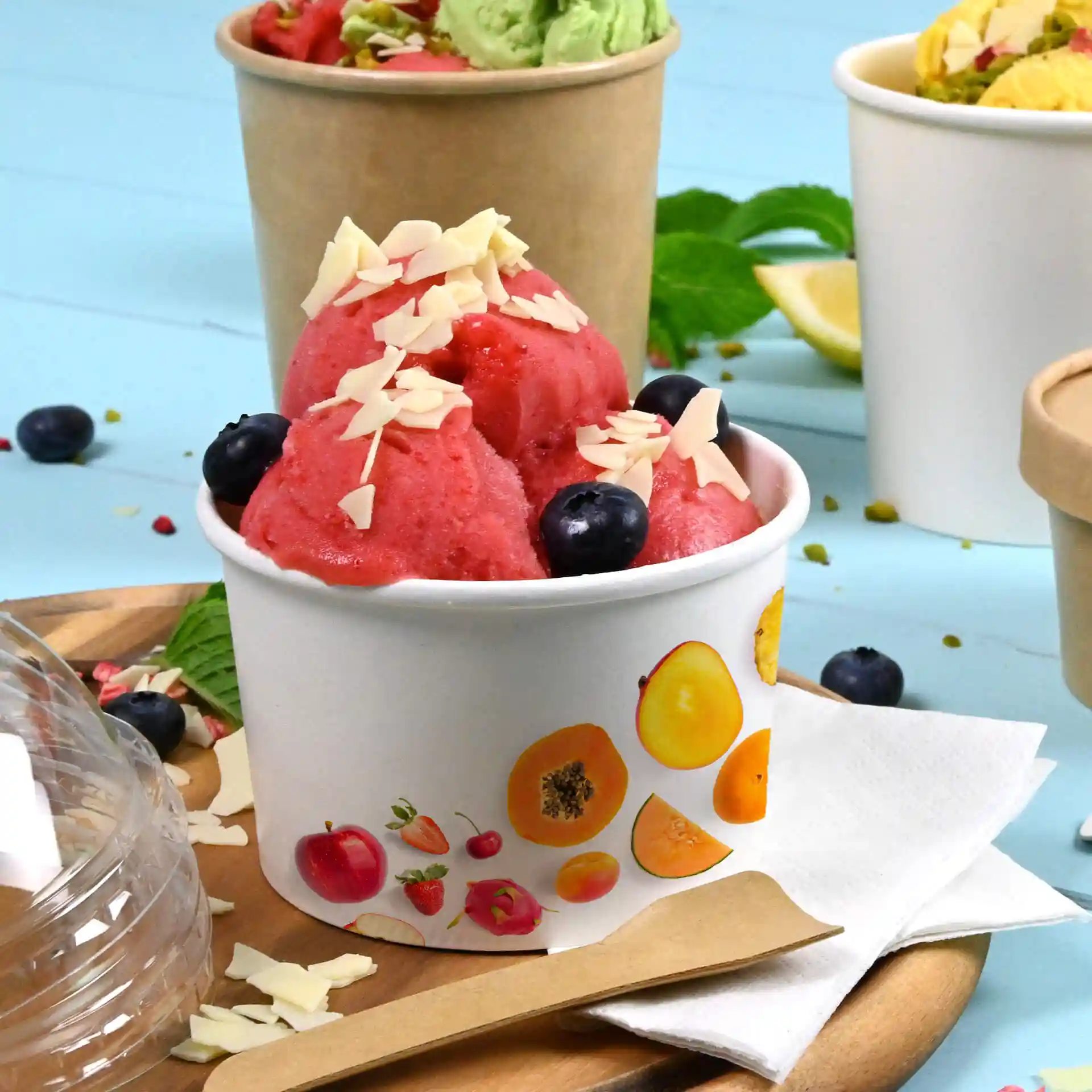 Coppa rotonda bianca di gelato riempita di sorbetto rosa, guarnita con scaglie di mandorle e mirtilli, su un vassoio di legno.