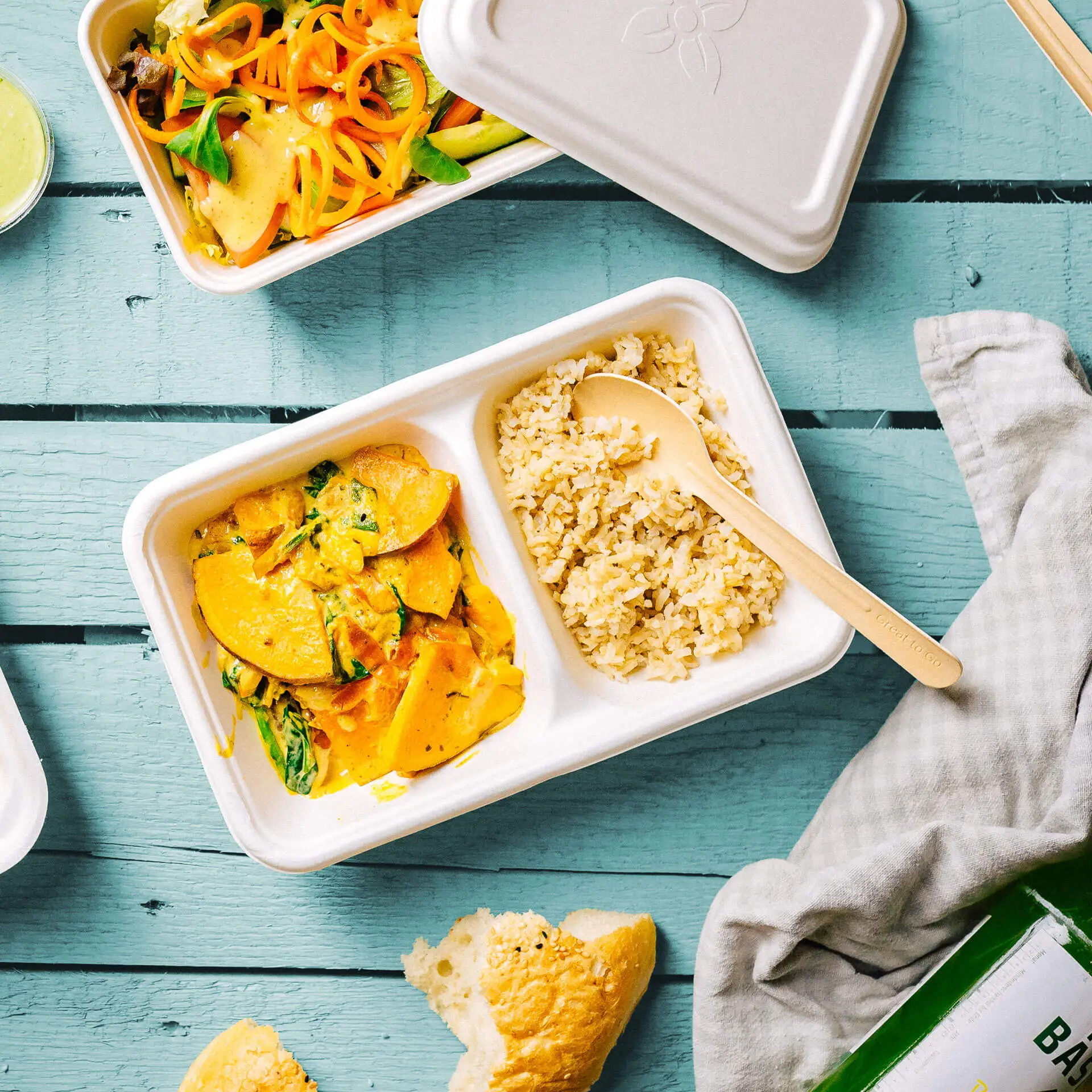 Una ciotola per menu biodegradabile in canna da zucchero con due scomparti; contiene un piatto di verdure giallo e riso marrone, con un cucchiaio di legno.