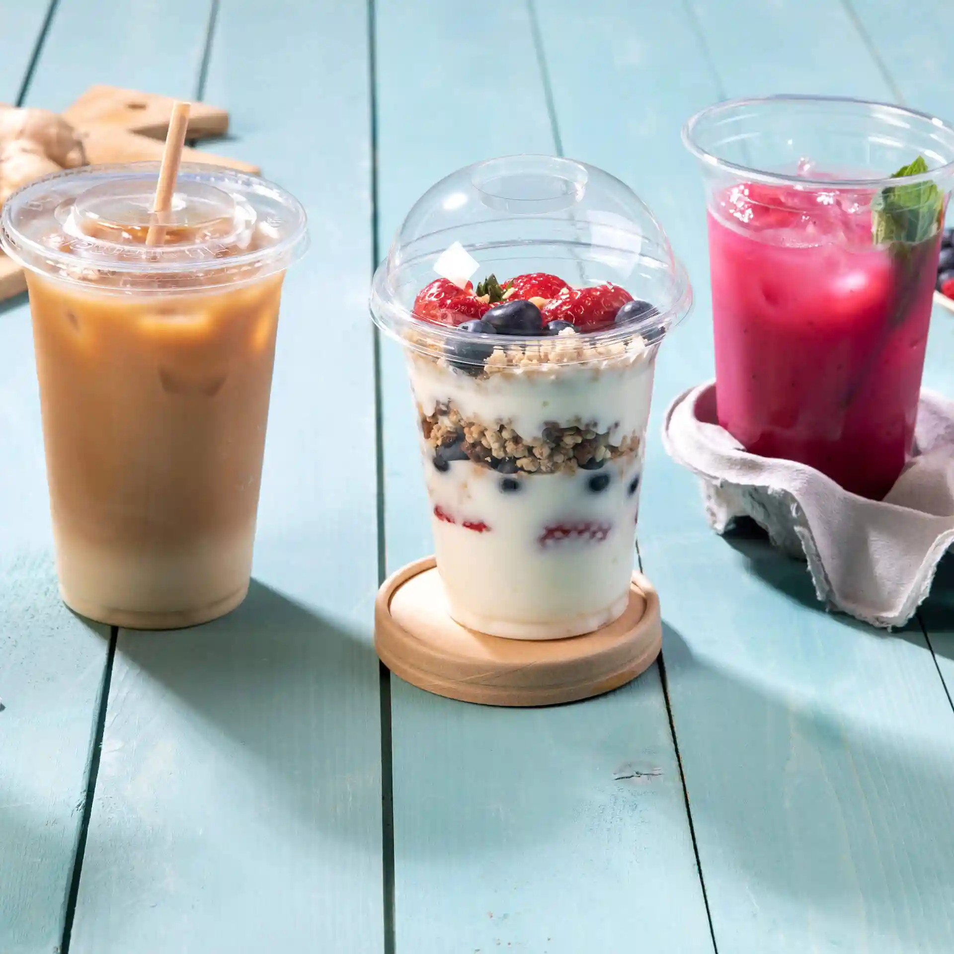 Immagine di tre bevande: caffè freddo, parfait di yogurt con frutti di bosco e una bevanda rosa su una superficie in legno blu.