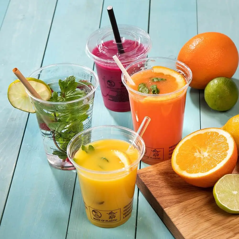 Quattro bicchieri di plastica trasparente riempiti di bevande colorate, tra cui succo d'arancia e uno smoothie rosa, accanto a frutta fresca.