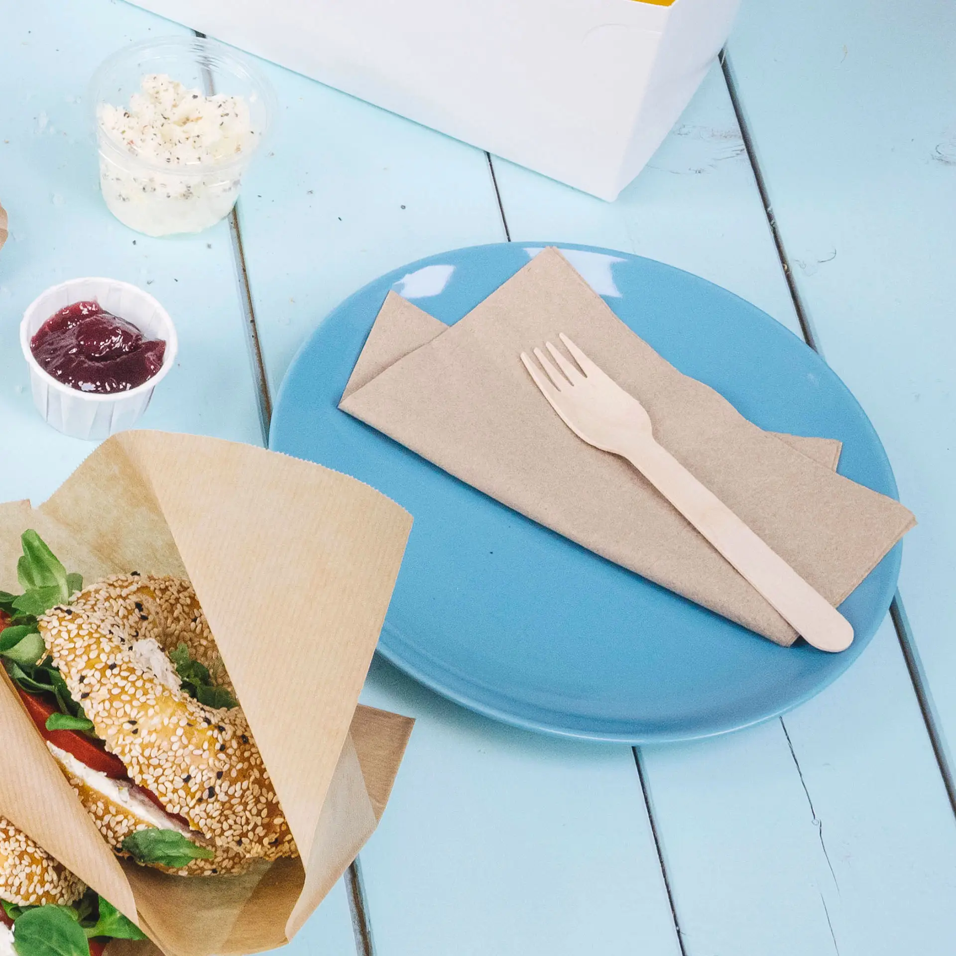 Piatto blu con tovagliolo marrone e forchetta di legno, accanto a un panino al sesamo e a un piccolo bicchiere di marmellata.