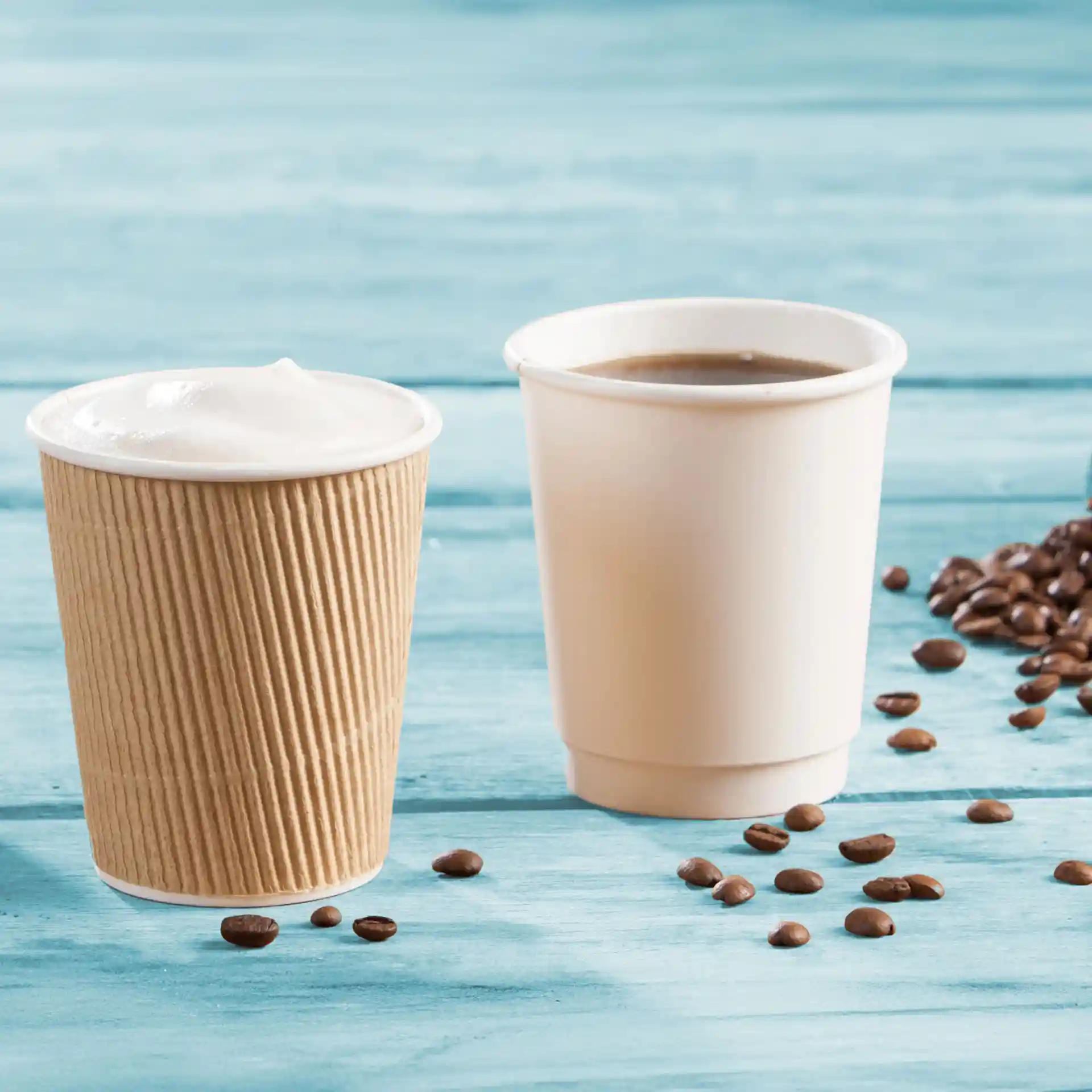 Bicchieri in cartone a doppia parete marroni e bianchi, con chicchi di caffè sparsi su una superficie in legno blu.