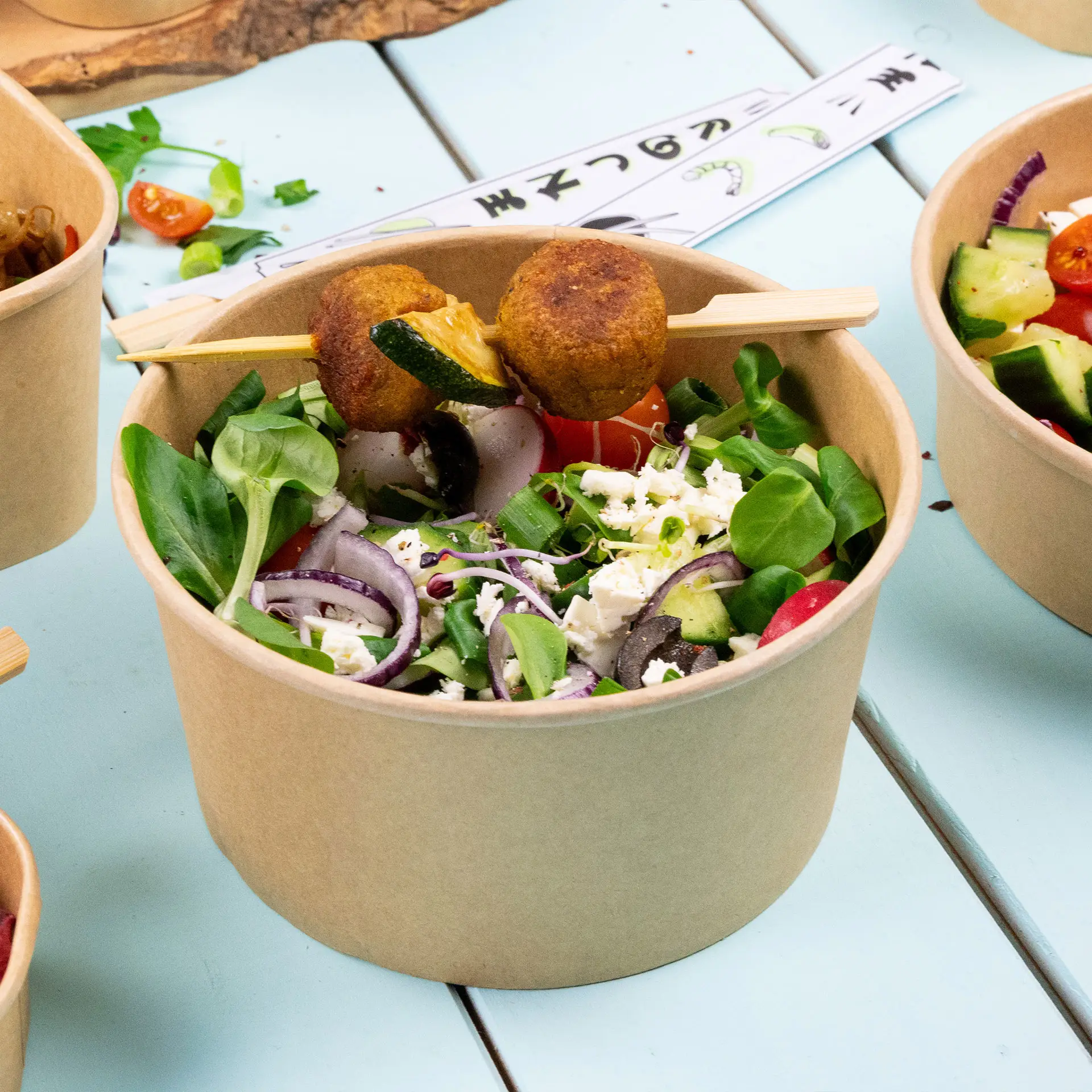 Spiedini in bambù che sorreggono cibo in porzioni mordi e fuggi su un'insalata in una ciotola marrone, circondati da ciotole simili su una superficie blu.