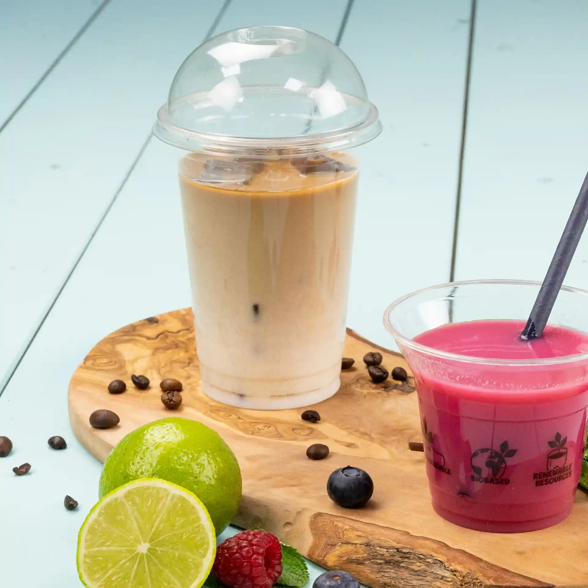 Immagine di un bicchiere di plastica trasparente da 16 oz riempito di caffè freddo, accanto a una bevanda rosa in un bicchiere trasparente su un tagliere di legno con frutta.