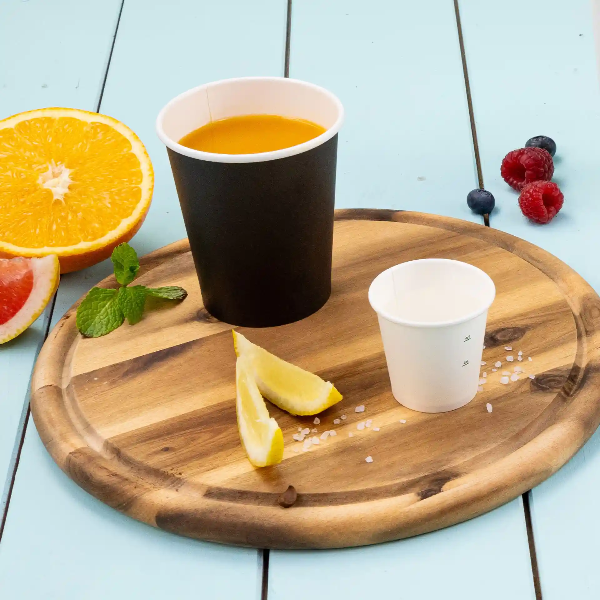 Bicchiere nero con liquido arancione, piccolo bicchiere bianco, spicchi di limone e frutta fresca su un vassoio di legno.