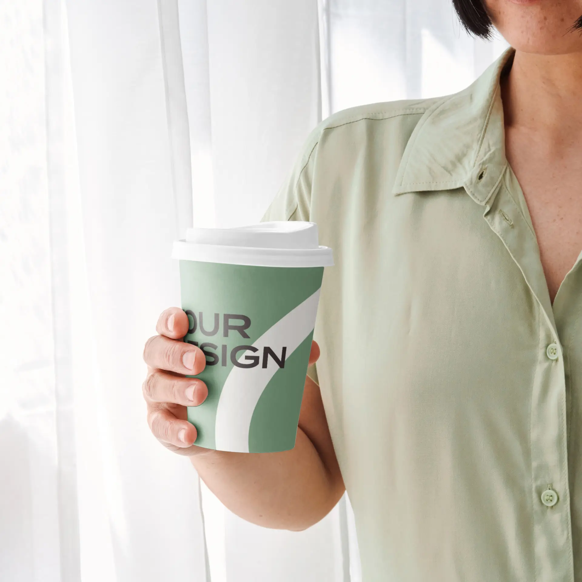 Bicchiere di carta verde a mano con coperchio bianco, con logo e design grafico; persona con una camicia verde chiaro sullo sfondo.