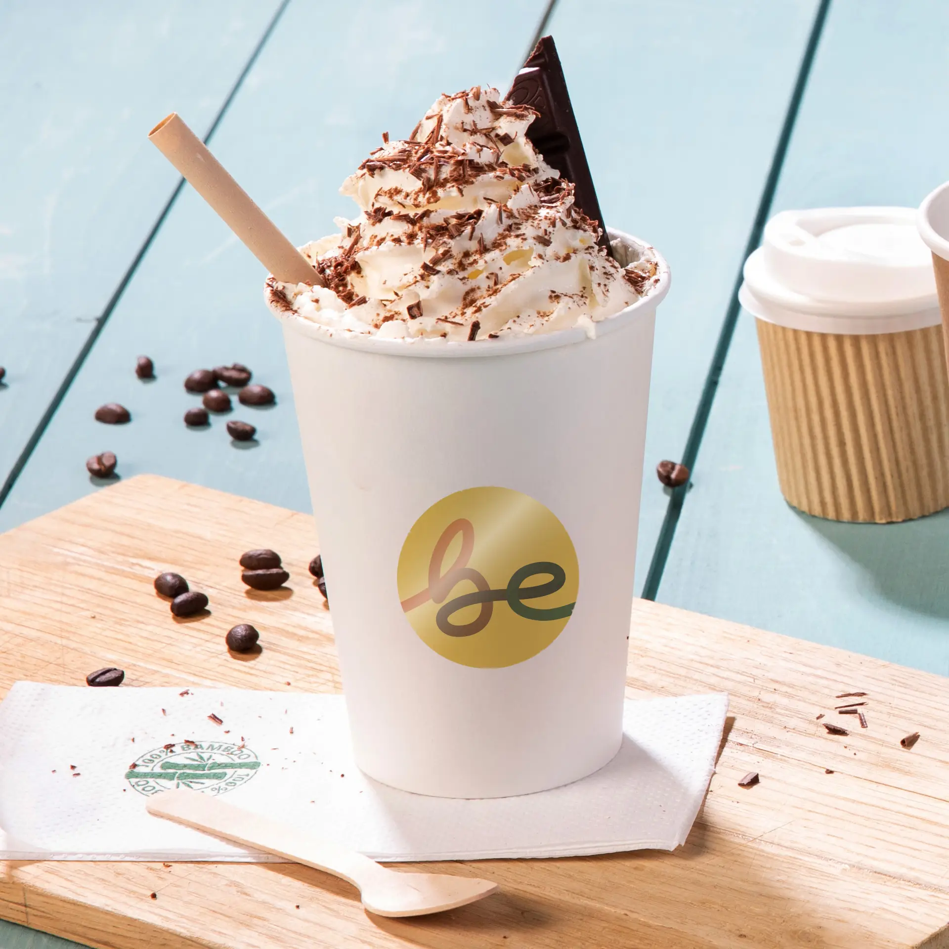 Becher con panna montata, scaglie di cioccolato, un pezzo di cioccolato e una cannuccia di carta, su una superficie di legno con chicchi di caffè.
