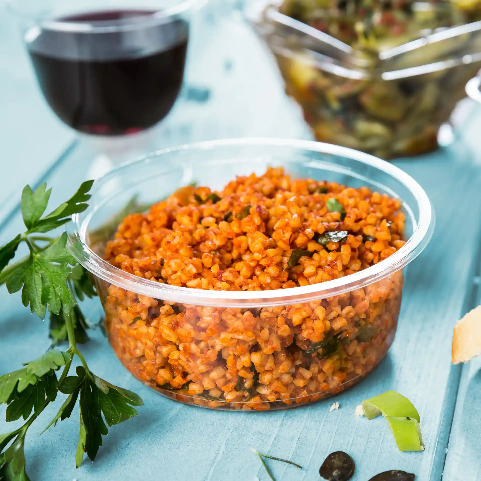 Contenitore deli trasparente rotondo riempito di bulgur arancione, guarnito con prezzemolo, su una superficie blu.