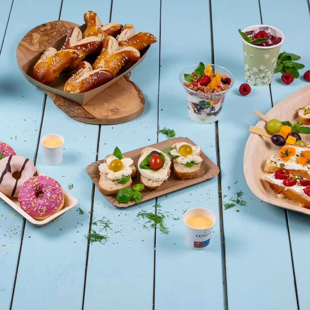 Una varietà di finger food tra cui pretzel, panini con condimenti, ciambelle e coppette di frutta su un tavolo di legno blu.