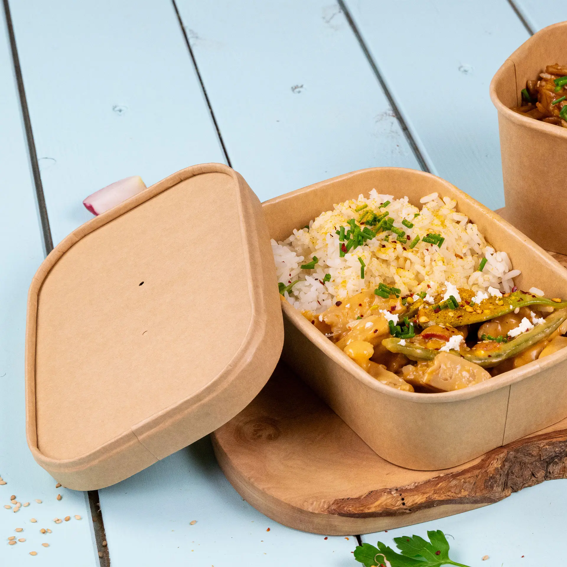 Contenitore in cartone con riso e verdure, parzialmente aperto; coperchio rotondo accanto su una superficie di legno.