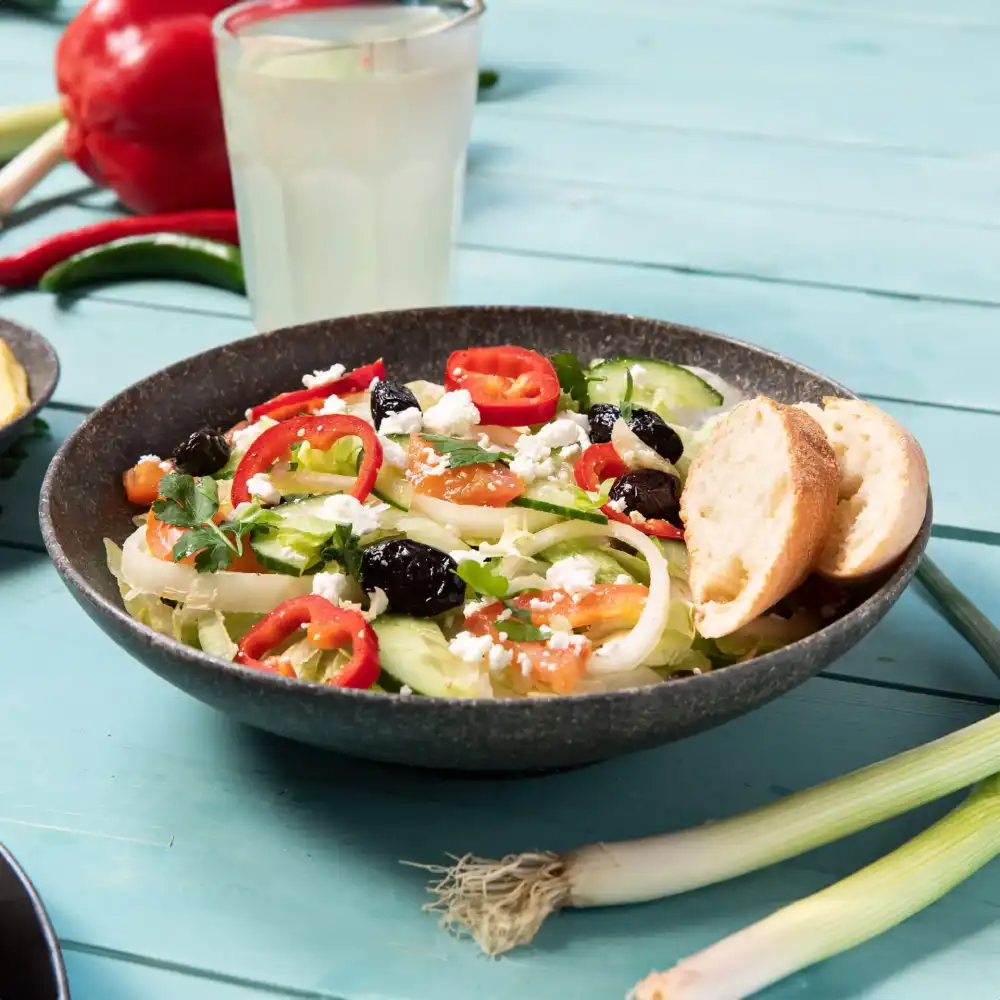 Ciotola di insalata con cetriolo, pomodori, olive, formaggio feta e fettine di peperone rosso, accompagnata da due fette di pane.