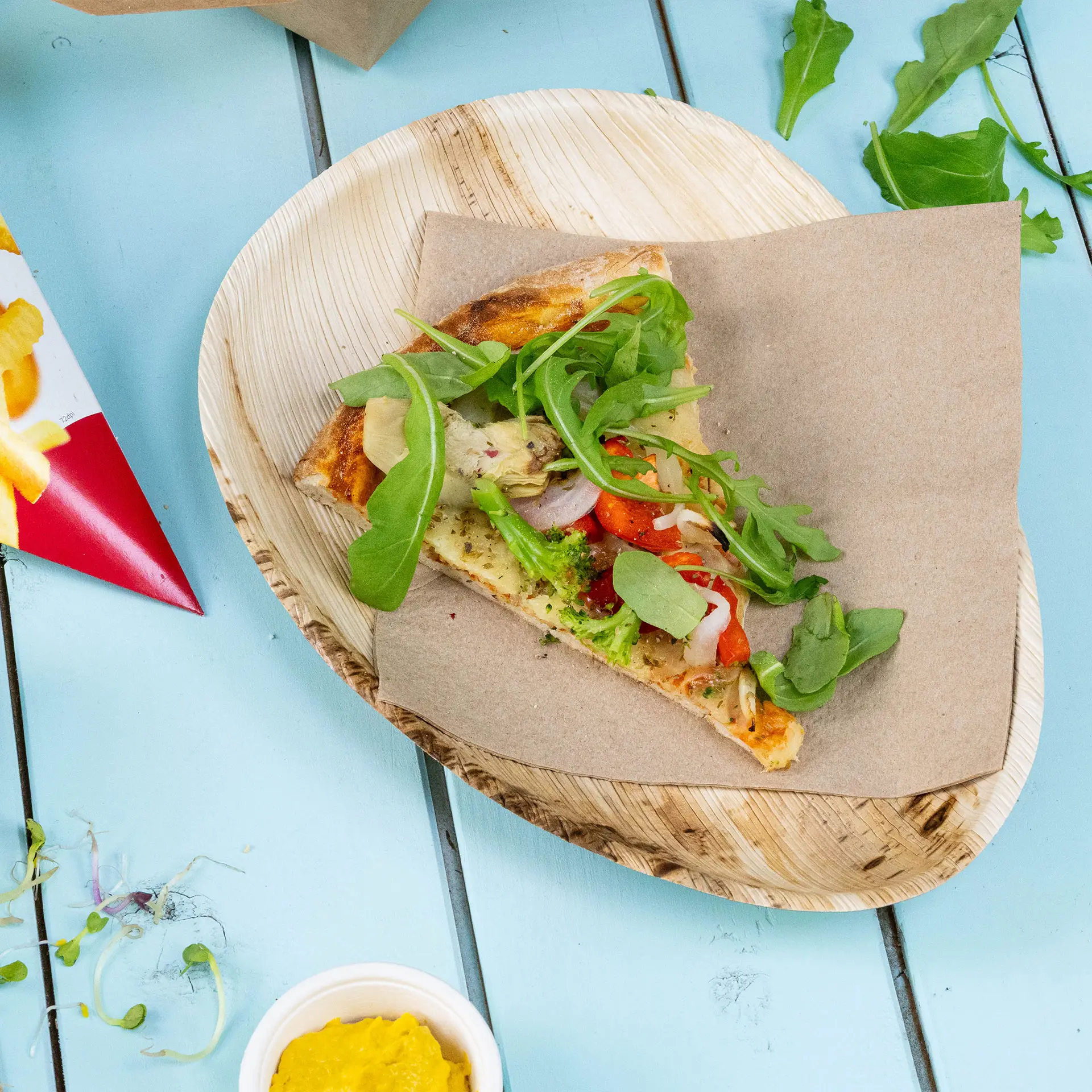Una fetta di pizza guarnita con verdure su un piatto a forma di goccia in foglia di palma; una piccola tazza di senape vicina.