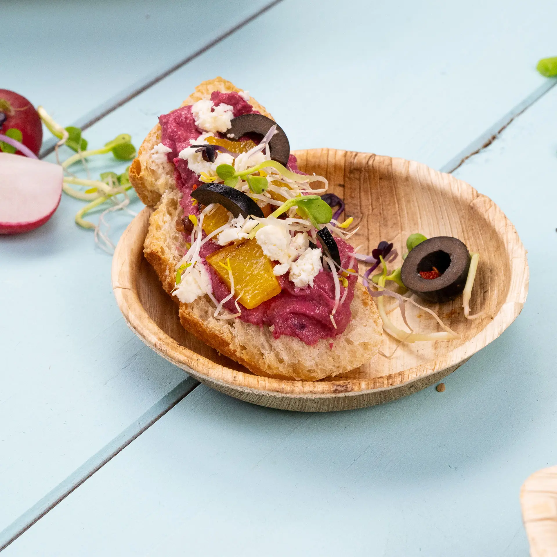 Mini ciotola in foglia di palma contenente una fetta di pane guarnita con una crema viola, olive, germogli e pezzi d'arancia su una superficie blu.