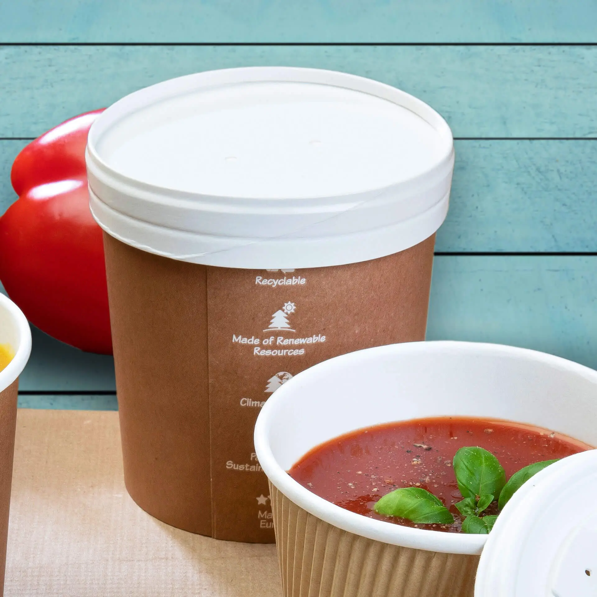 Bicchiere di cartone per zuppa con coperchio in membrana bianca, rivestito in bio-PE; guarnizione di basilico in un bicchiere vicino; pomodoro rosso sullo sfondo.