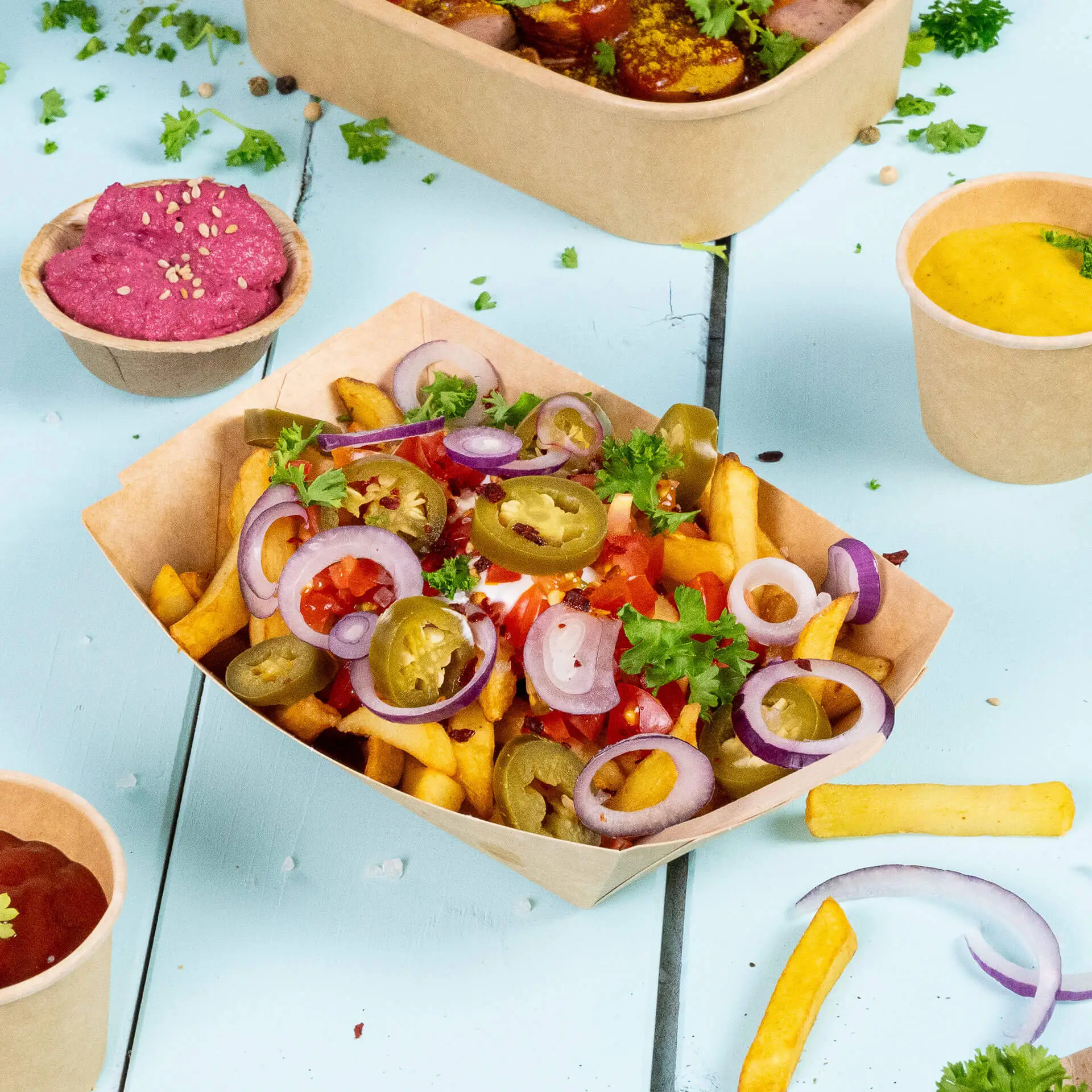 Un vassoio di cartone marrone per snack riempito di patatine guarnite con pomodori, jalapeños, cipolle ed erbe; vassoi e salse aggiuntive sullo sfondo.