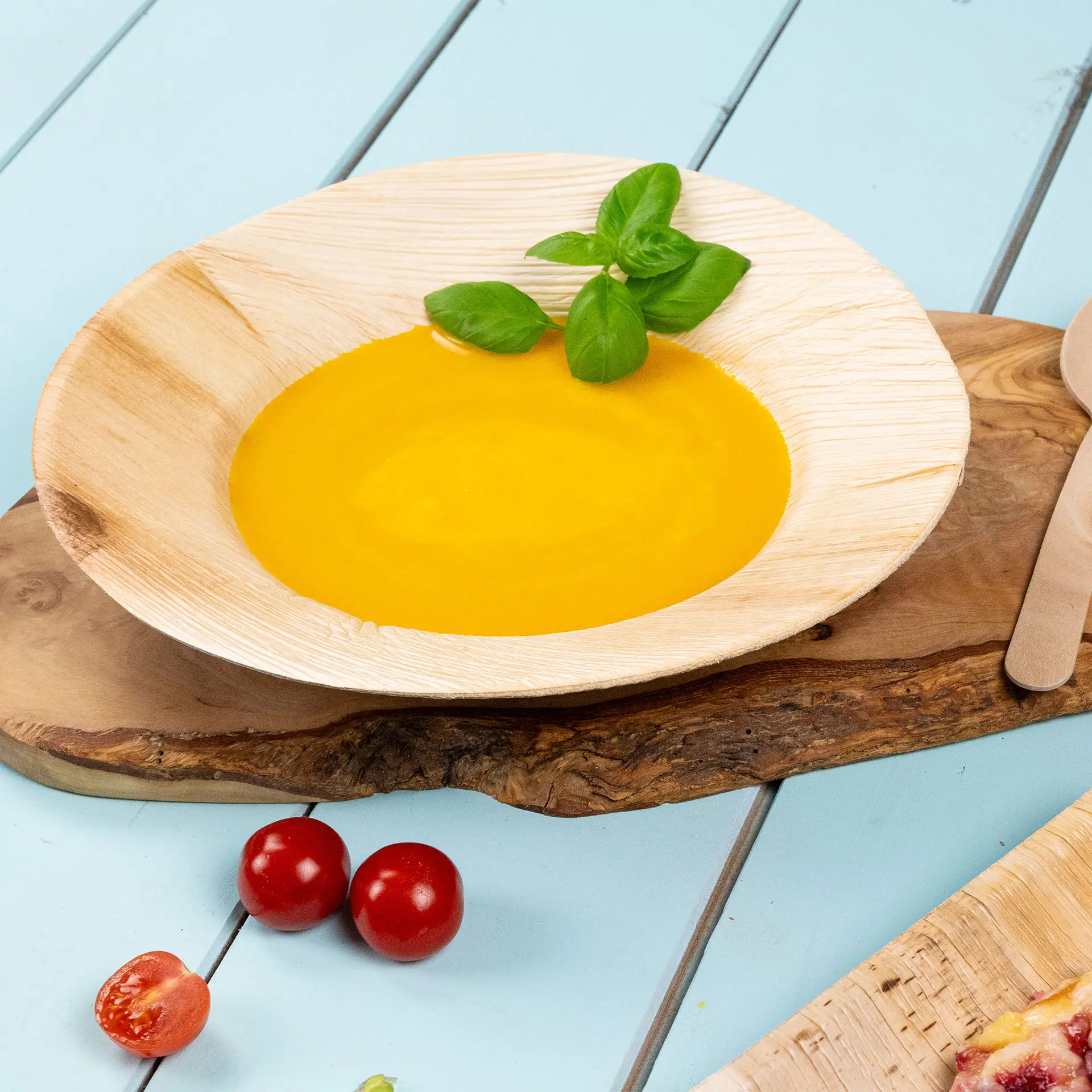 Un piatto in foglia di palma, diametro 23 cm, riempito con zuppa dorata e guarnito con foglie di basilico; pomodorini nelle vicinanze.