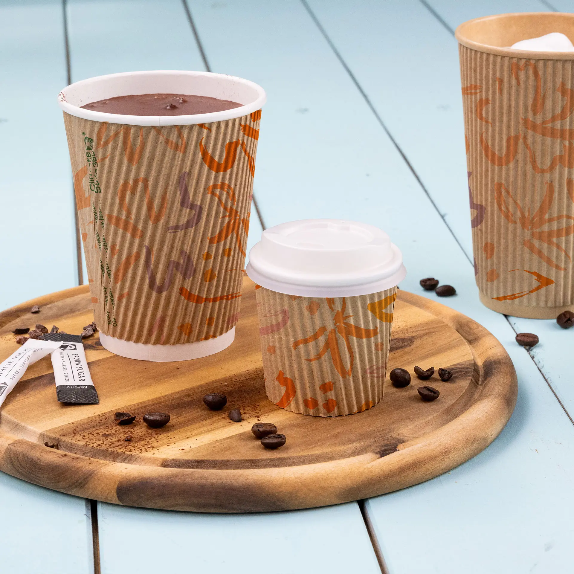 Bicchieri da caffè marroni rigati su un vassoio di legno, con un grande bicchiere di bevanda calda, un bicchiere più piccolo con coperchio e chicchi di caffè.