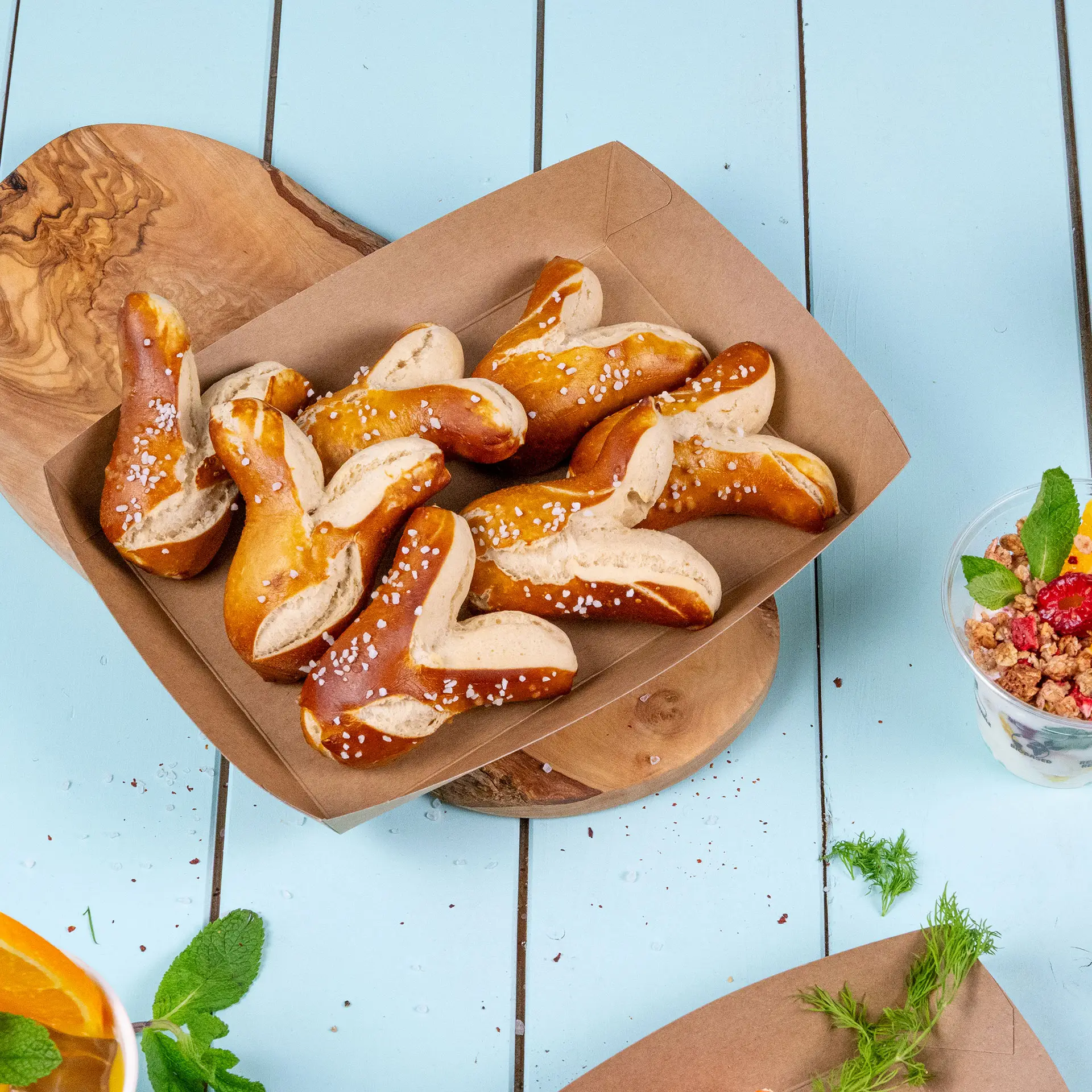 Un vassoio di cartone marrone ripieno di pretzel, cosparso di sale grosso, posato su un tagliere di legno.