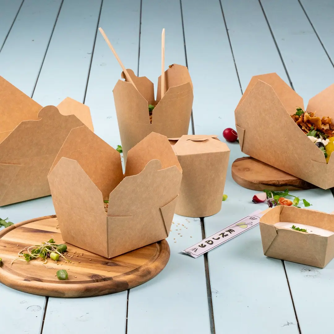 Contenitori alimentari in cartone marrone di varie forme su un tagliere di legno con ingredienti freschi sparsi.