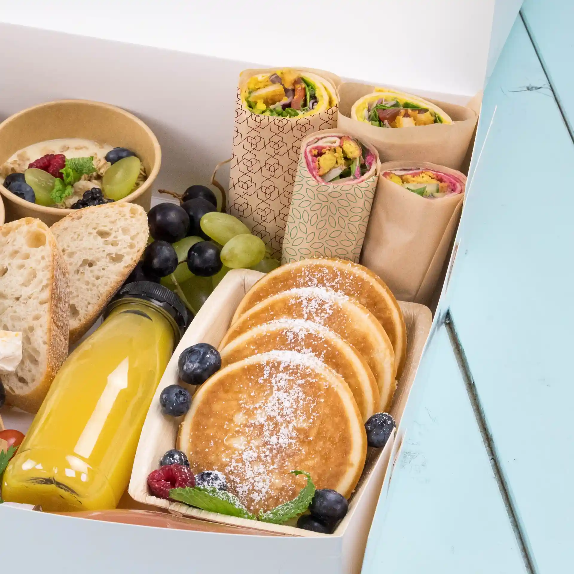 Varietà di alimenti tra cui wrap, pancake, frutta e una bevanda in una scatola su una superficie blu.