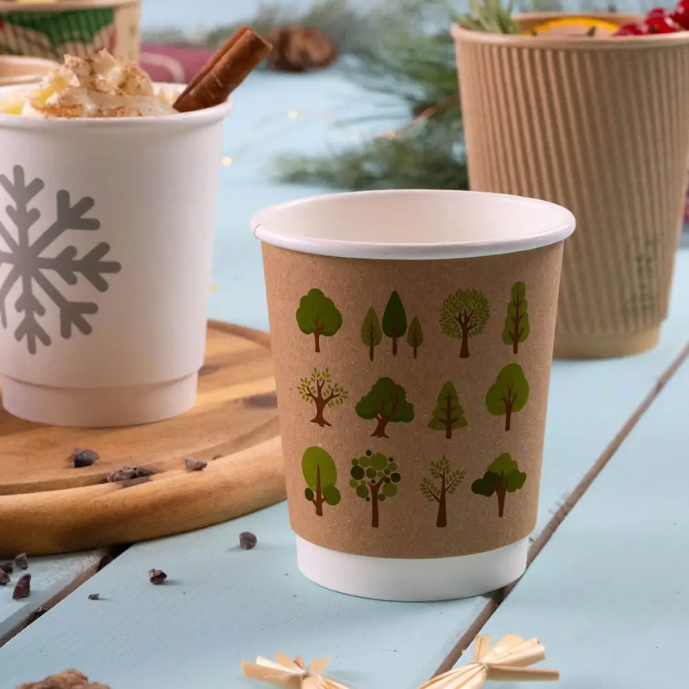 Becher in carta kraft con grafiche di alberi e piante verdi, posato su una superficie di legno con altri bicchieri sullo sfondo.