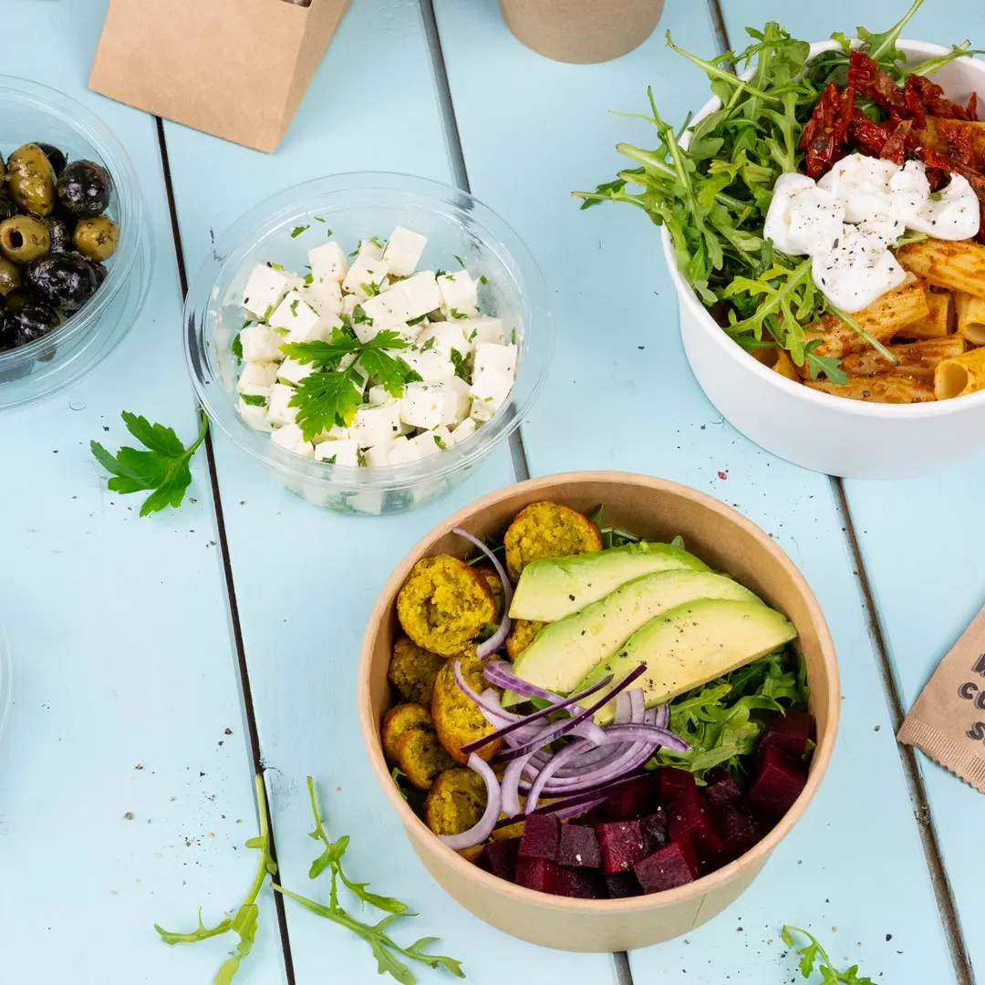 Ciotola con falafel, avocado, barbabietola, cipolla rossa e verdure, accanto a contenitori di feta e olive su una superficie blu.