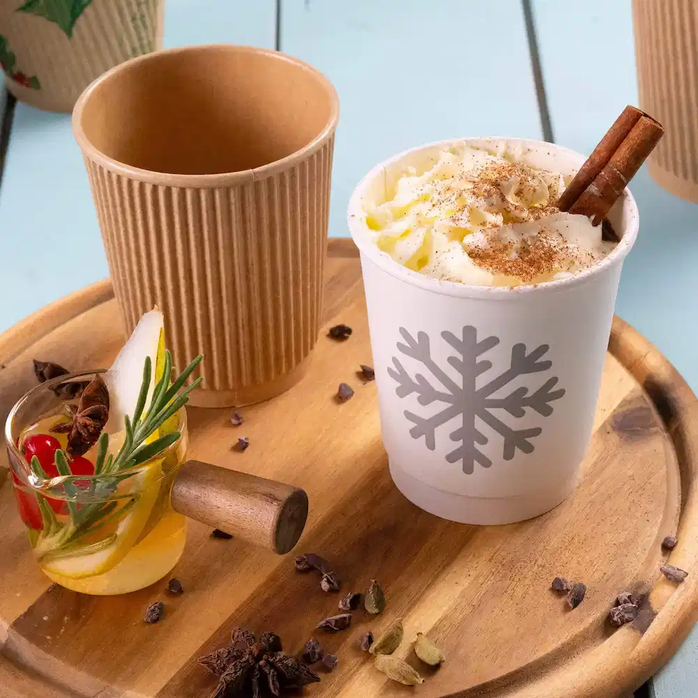 Immagine di un bicchiere di carta bianco a doppia parete con un design di fiocchi di neve, riempito di panna montata, accanto a un bicchiere di carta marrone rigato e a un bicchiere decorato.