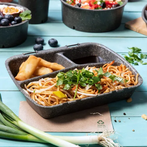 Scatola di pasti con noodles, guarnizione di verdure e involtini primavera, posizionata su un tavolo di legno blu con ingredienti freschi attorno.